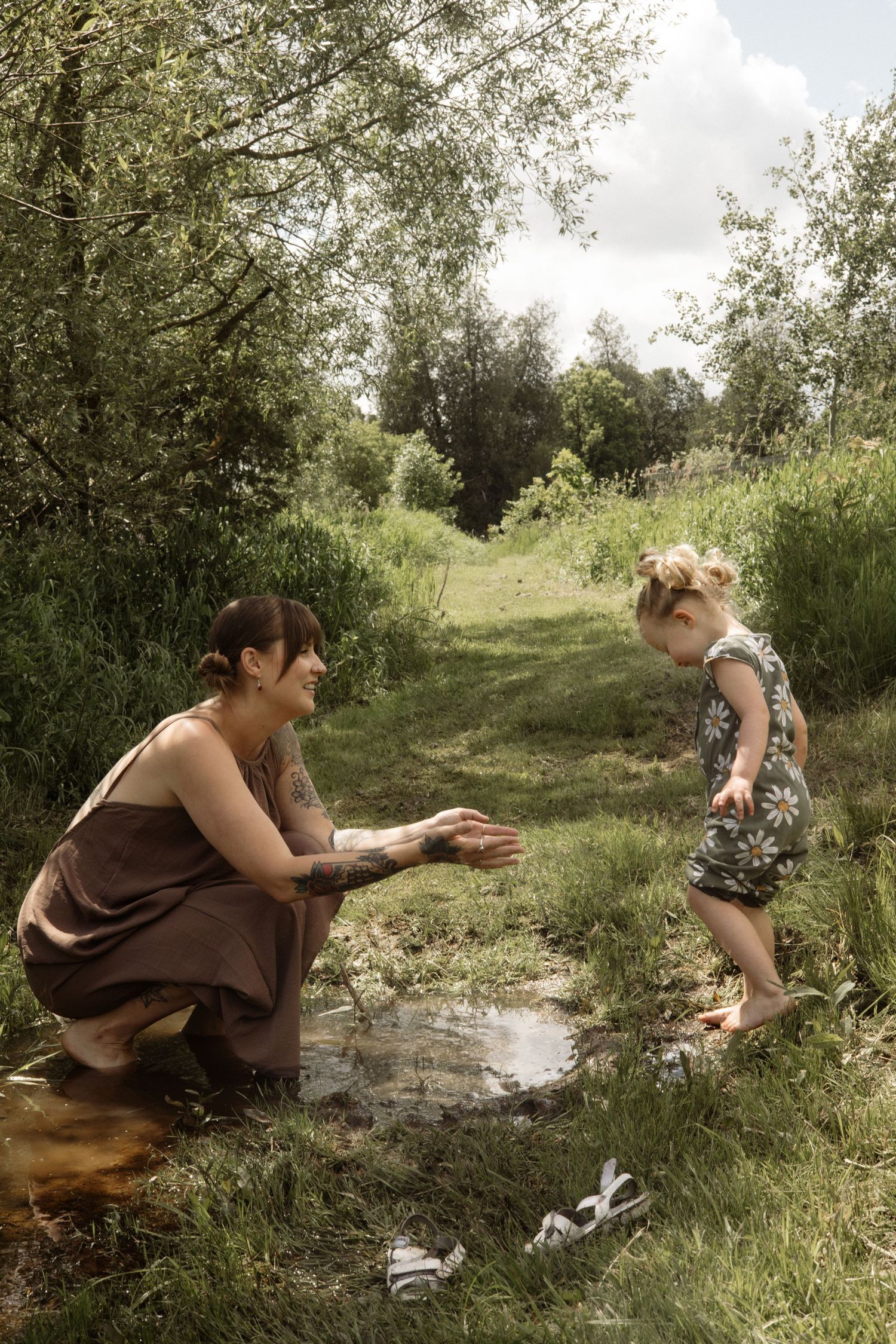 Mother kneeling by a muddy puddle reaching out to her barefoot toddler daughter in a floral romper outdoors.