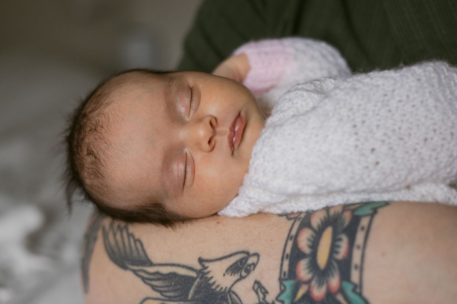 Sleeping newborn baby wrapped in white swaddle resting on tattooed arm.