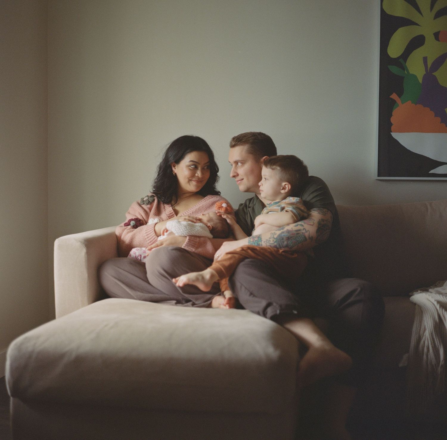 Young family with newborn and toddler cuddles together on beige sofa in cozy home.