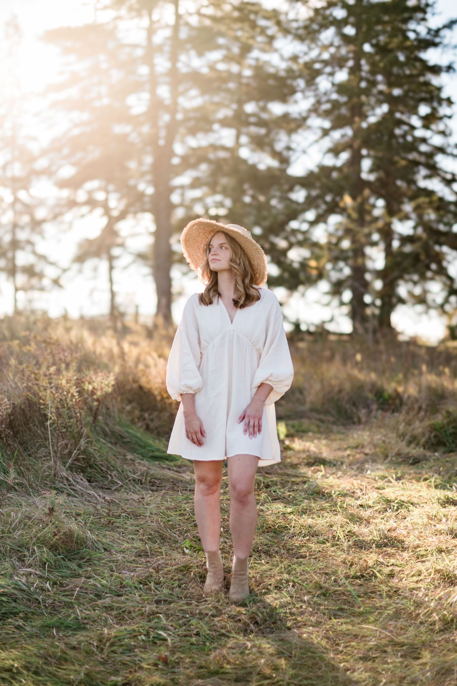 Woman in white dress and straw hat standing in sunlit forest path.