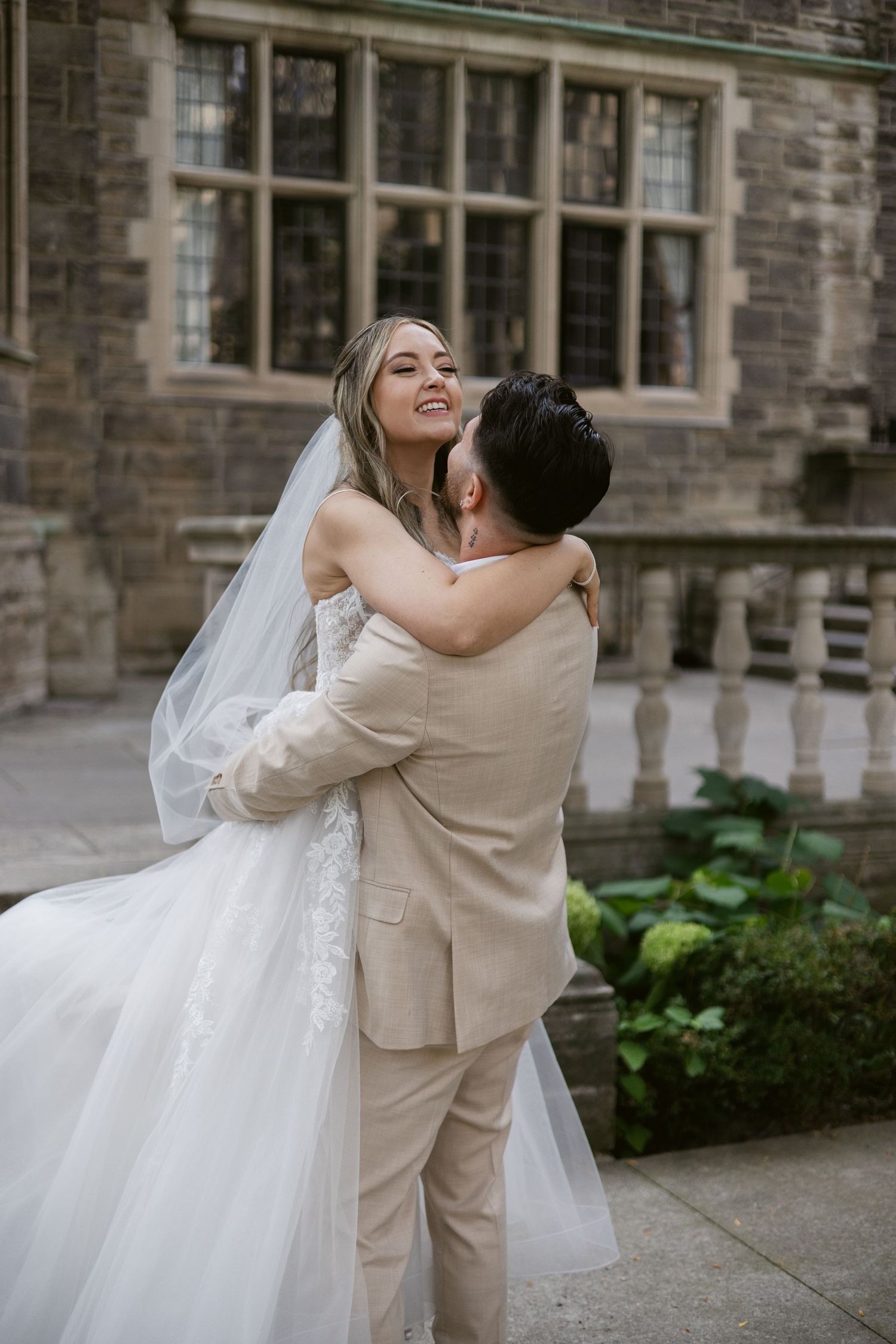 Groom in beige suit lifts smiling bride in white lace gown and veil outside stone building.