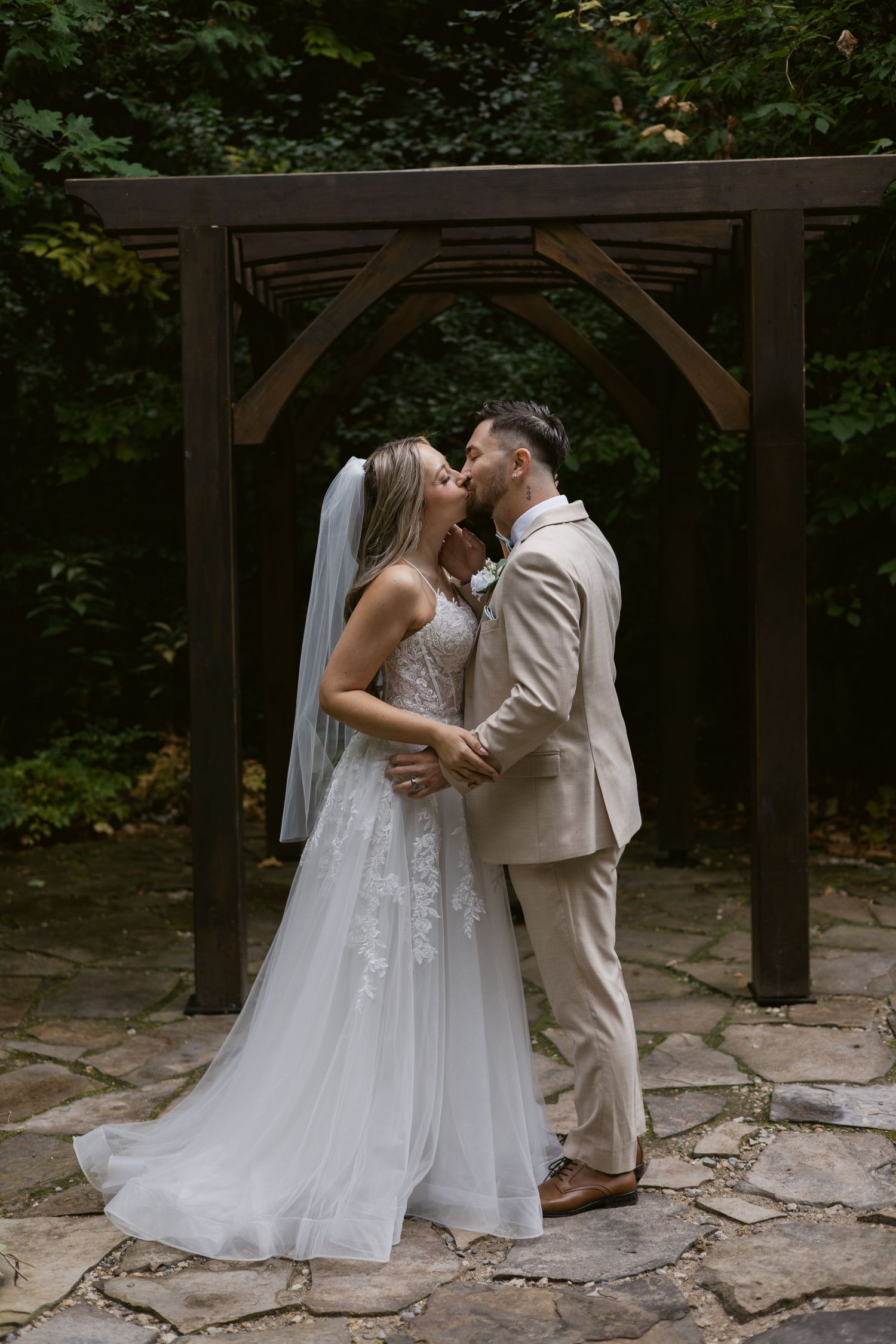 Newlyweds kissing under wooden pergola arch on stone patio with green forest backdrop.