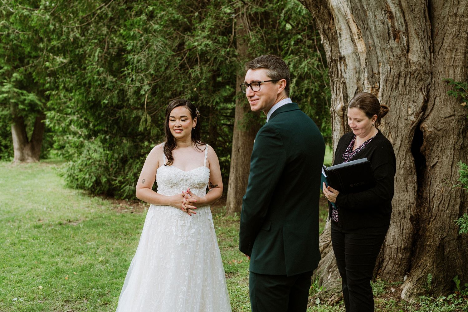 Bride and groom laugh joyfully with officiant during outdoor wedding ceremony beside a large tree.