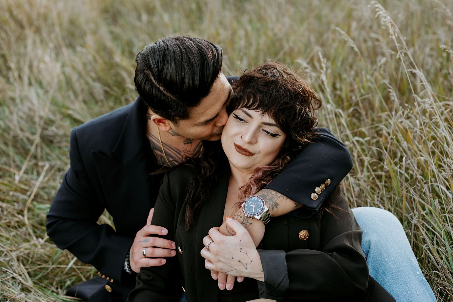 Couple sitting in tall grass field, man in black blazer embracing woman with curly hair during autumn engagement session.