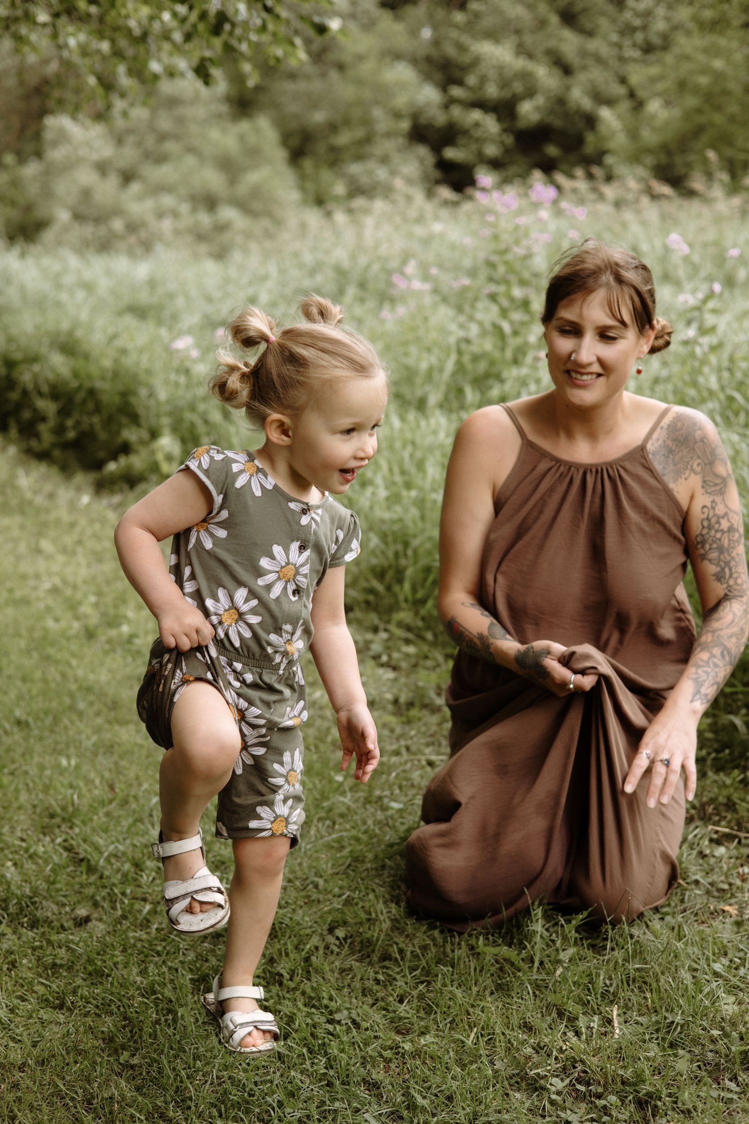 A smiling tattooed mother kneels on grass watching her happy young daughter run in a floral romper outdoors.
