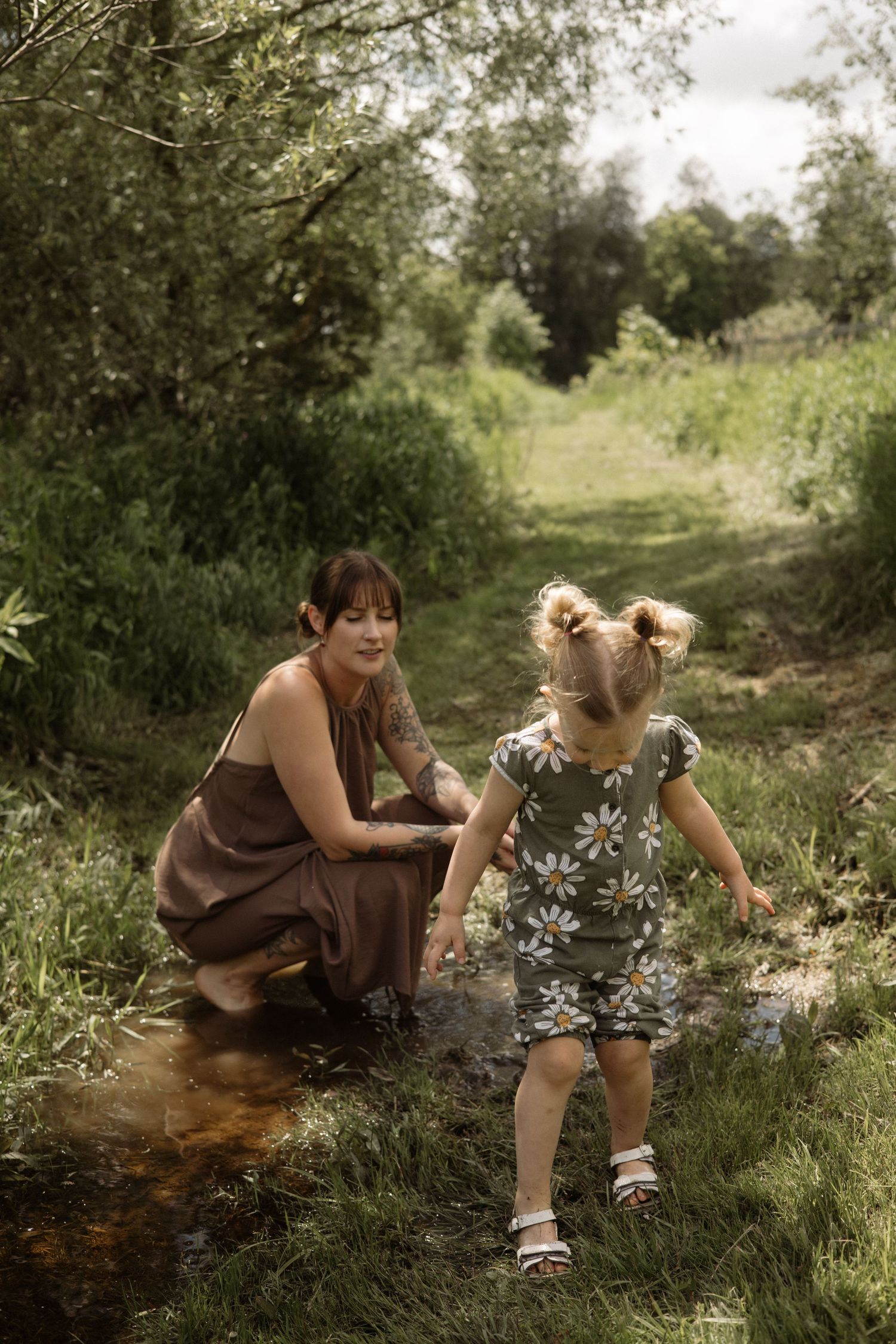 Tattooed mother crouching beside young daughter in daisy print outfit playing in a small stream surrounded by green nature.