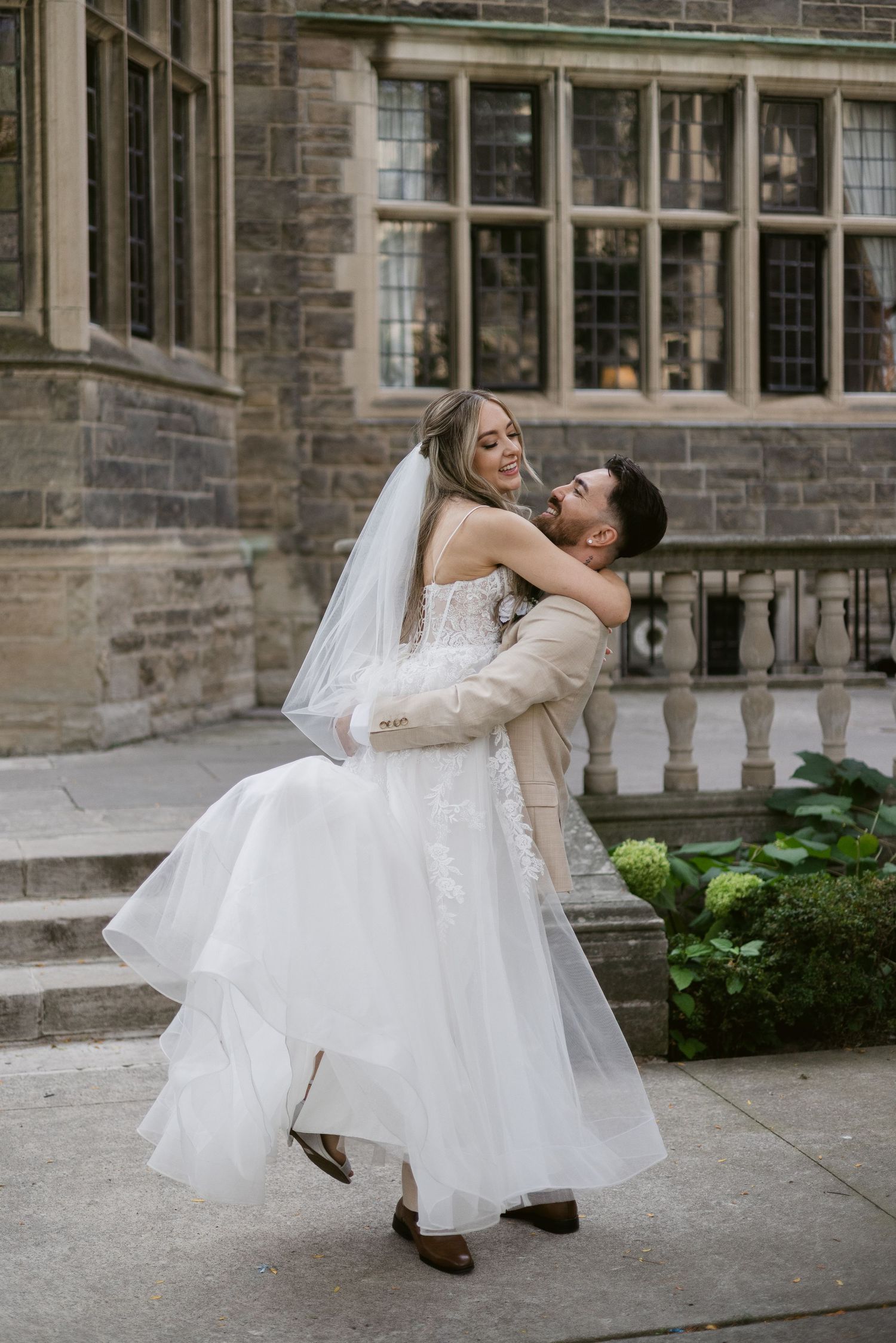 Groom sweeps bride off her feet in a romantic lift outside a historic stone building.