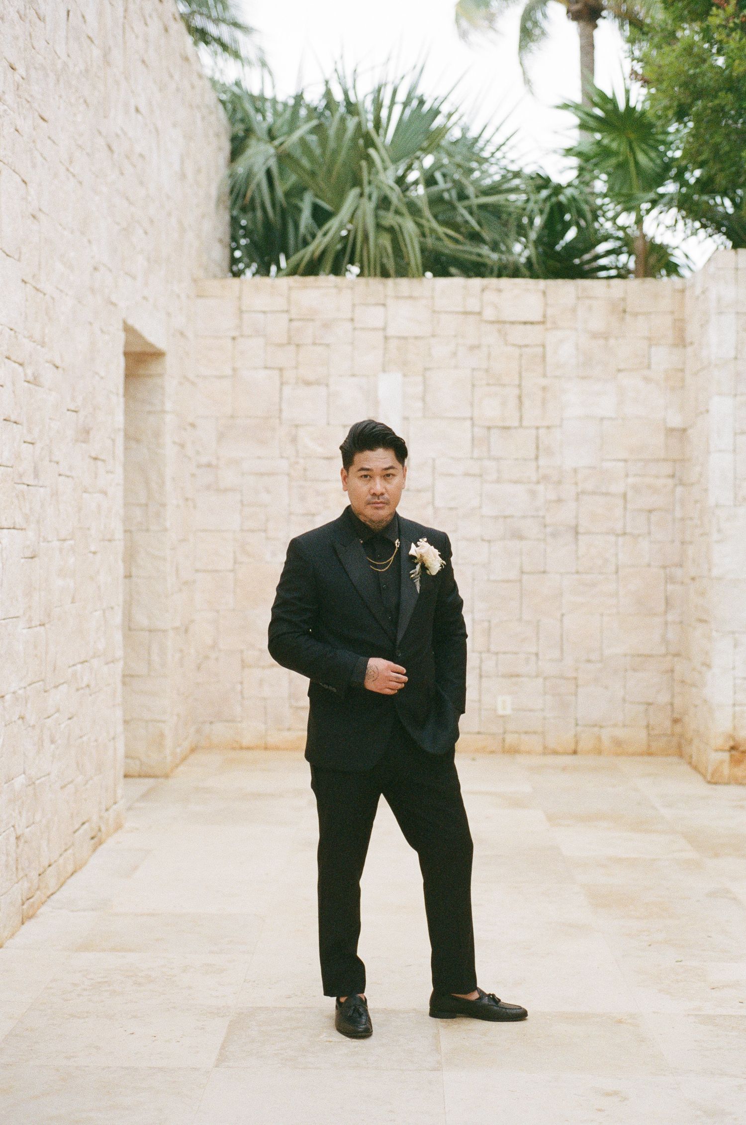 Groom in black suit with white boutonniere standing in stone courtyard with tropical palms.