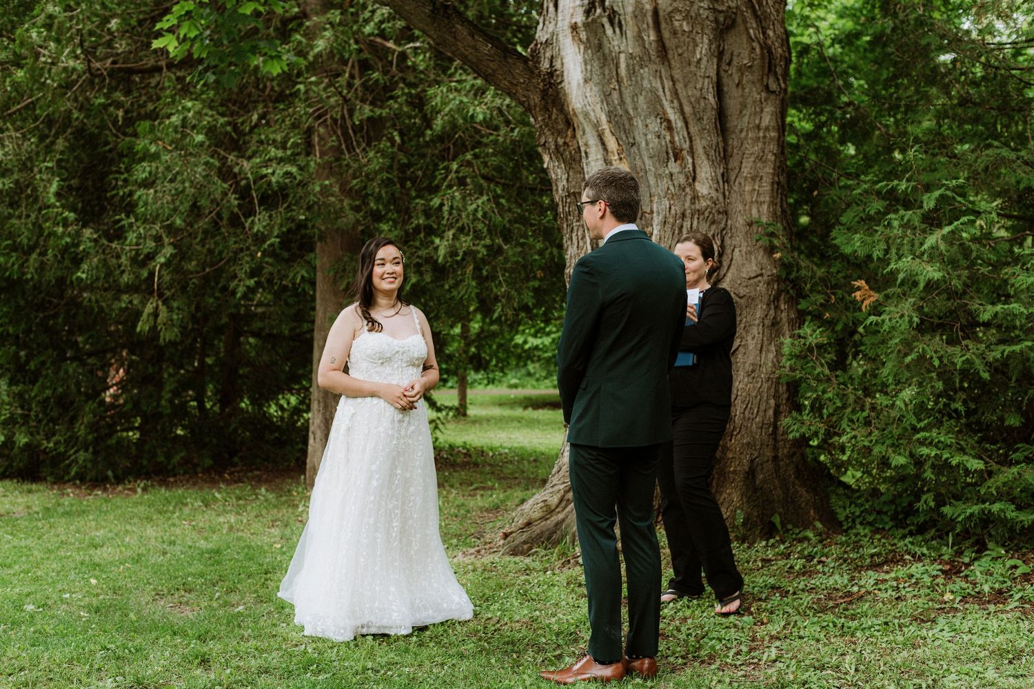 Bride smiles at groom during intimate outdoor wedding ceremony beside a large tree with officiant.