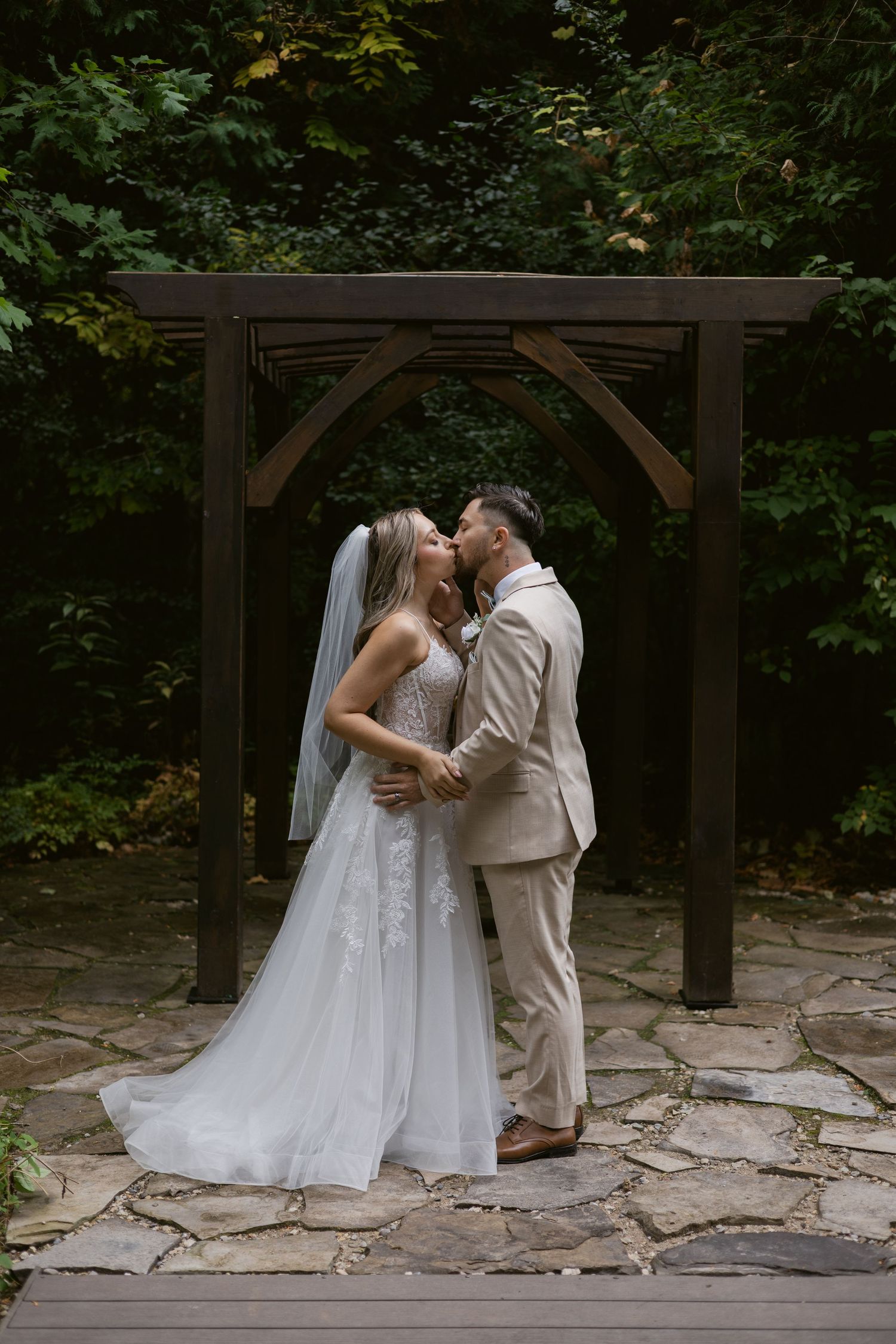 Bride and groom sharing first kiss under rustic wooden arch surrounded by lush green trees.
