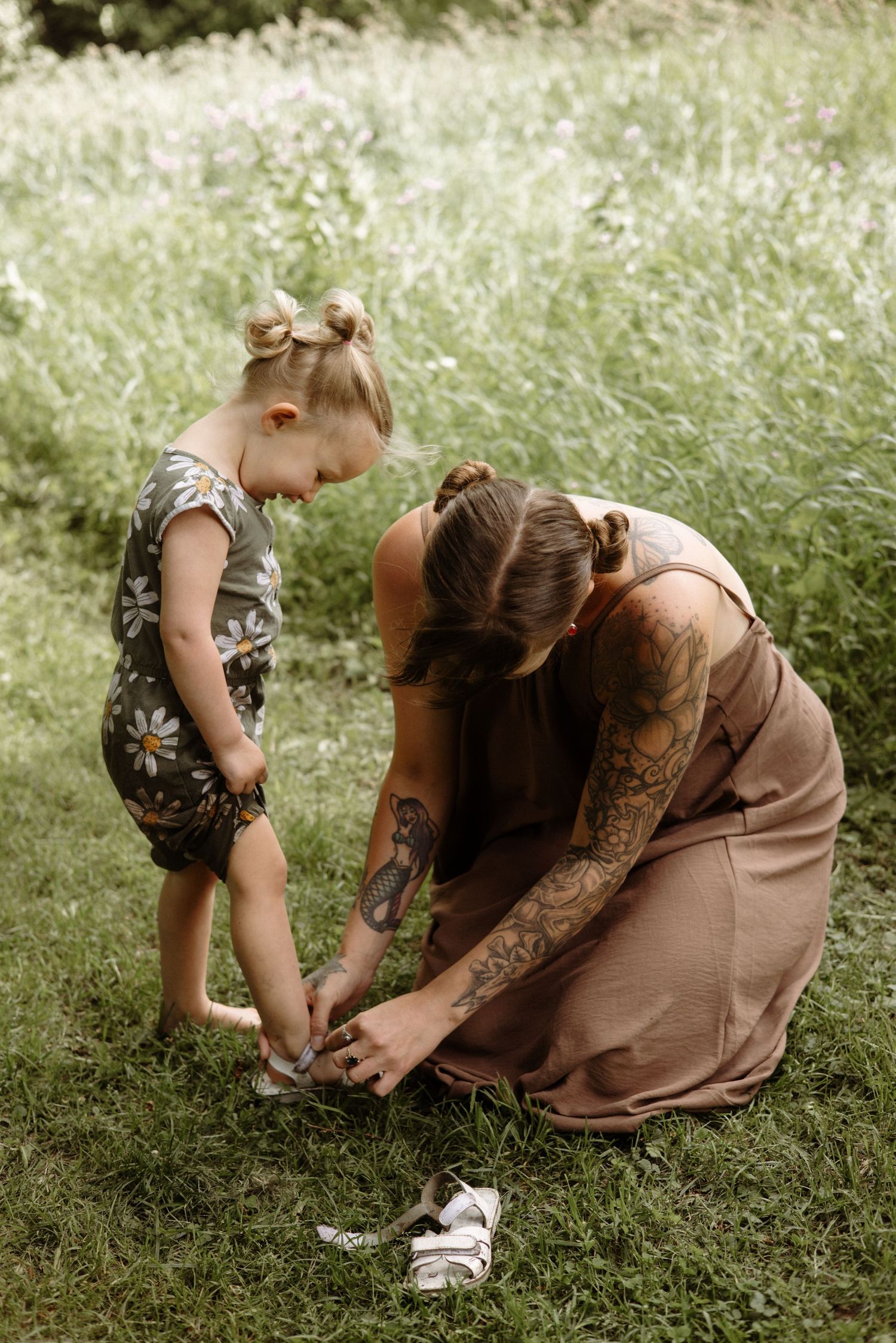 A tattooed mother crouches on green grass to fasten her toddler daughters shoes in a natural outdoor setting.