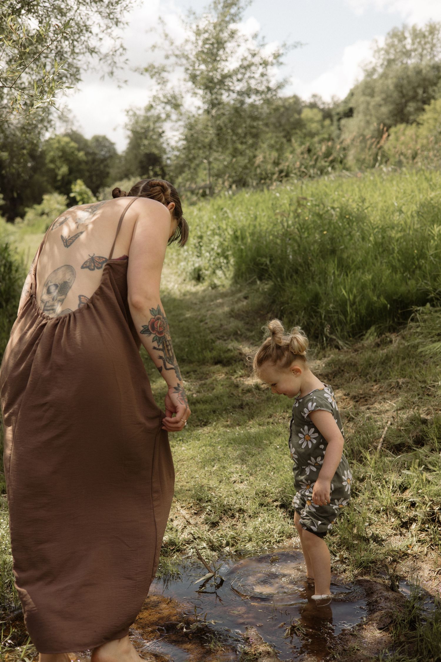 Tattooed mother in brown dress watching young daughter in daisy outfit explore a shallow stream in lush greenery.