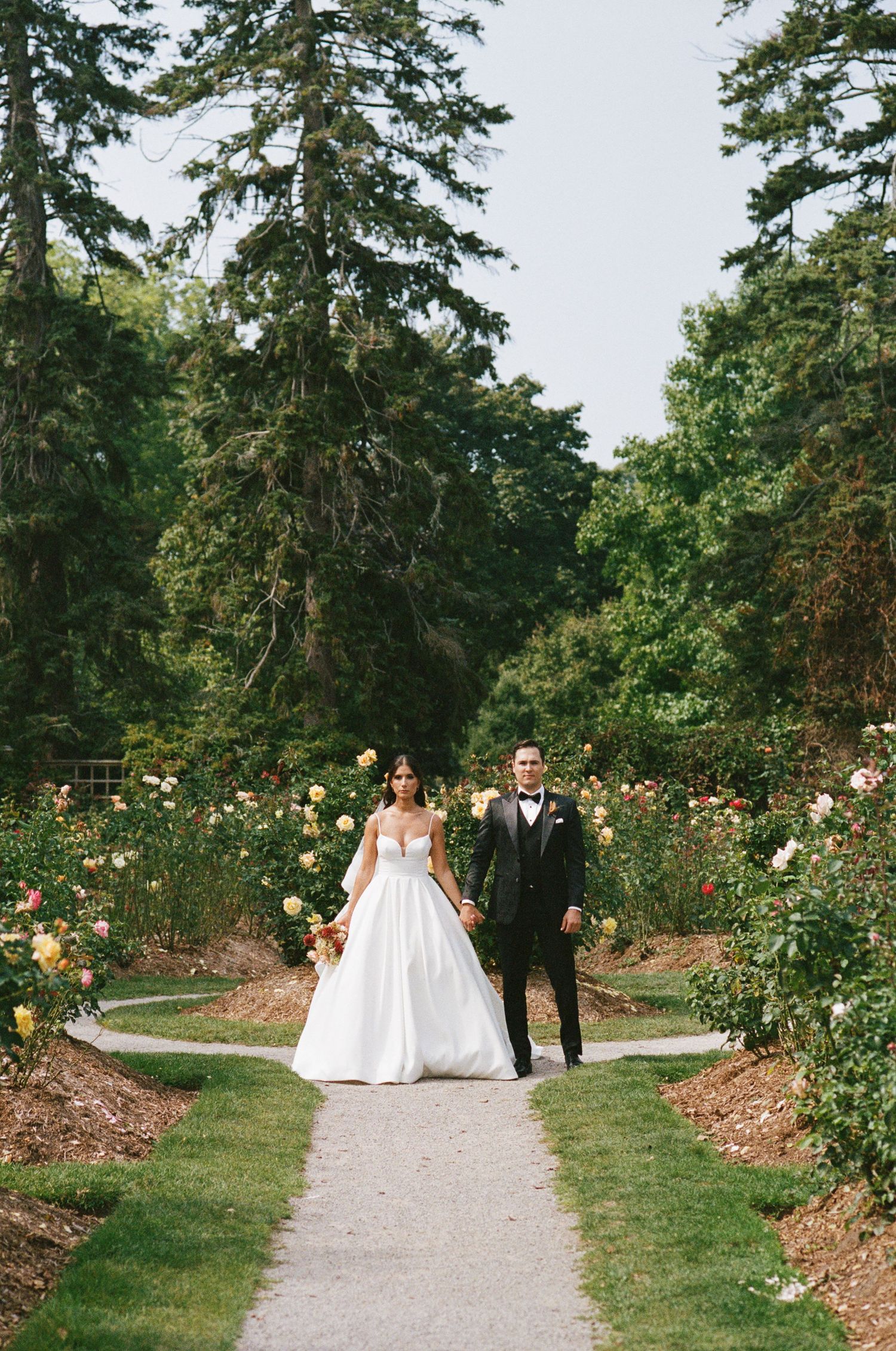 Bride and groom walk along paved garden path framed by tall trees and vibrant rose gardens.