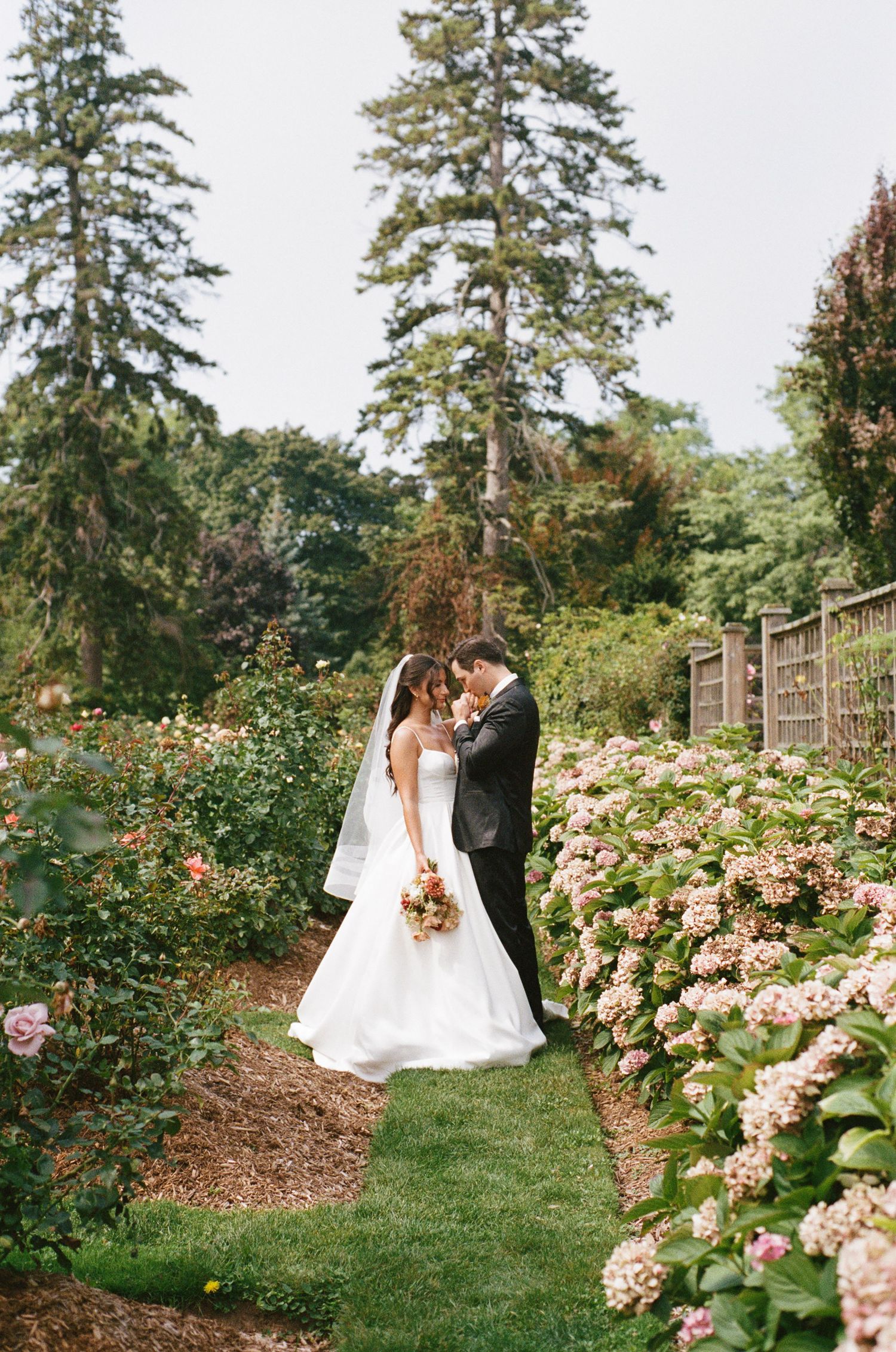 Wedding couple embracing on a garden path flanked by colorful roses and pink hydrangeas in bloom.