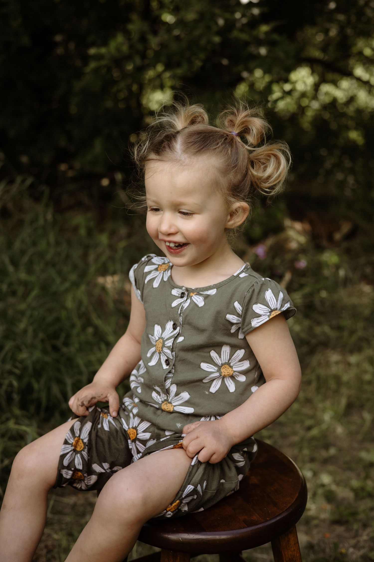 Happy toddler girl with pigtail buns laughing while sitting on wooden stool wearing olive green daisy print romper.