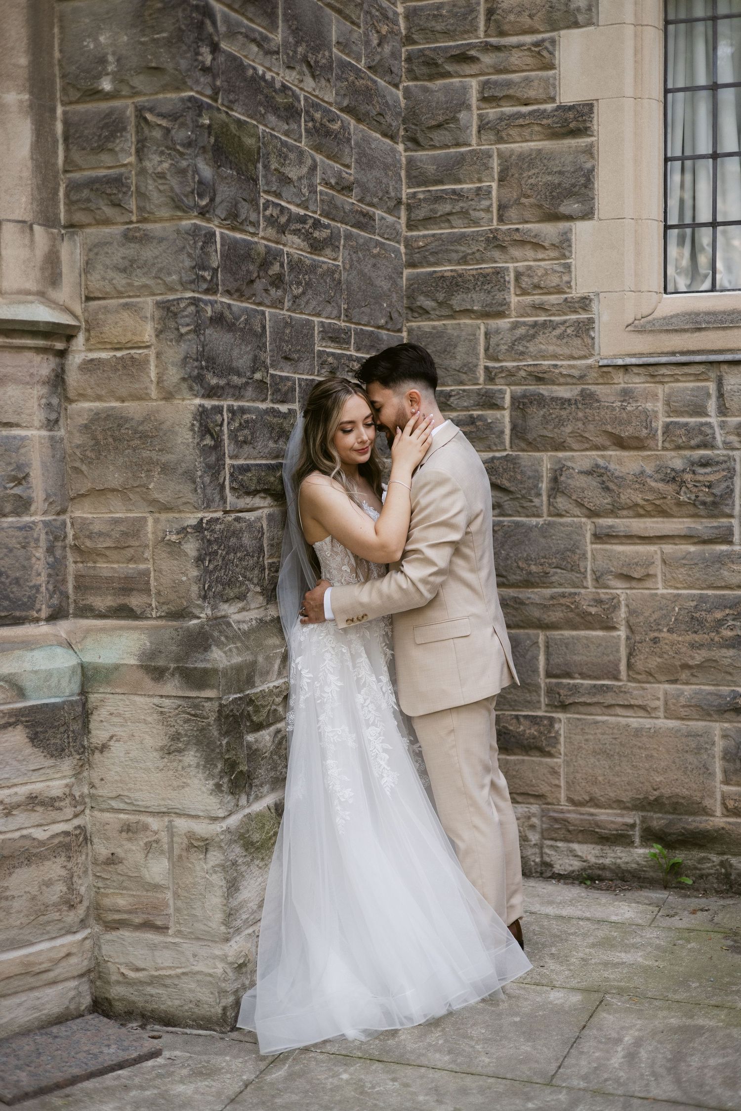 Bride in lace gown and groom in beige suit embrace tenderly against a rustic stone wall corner.