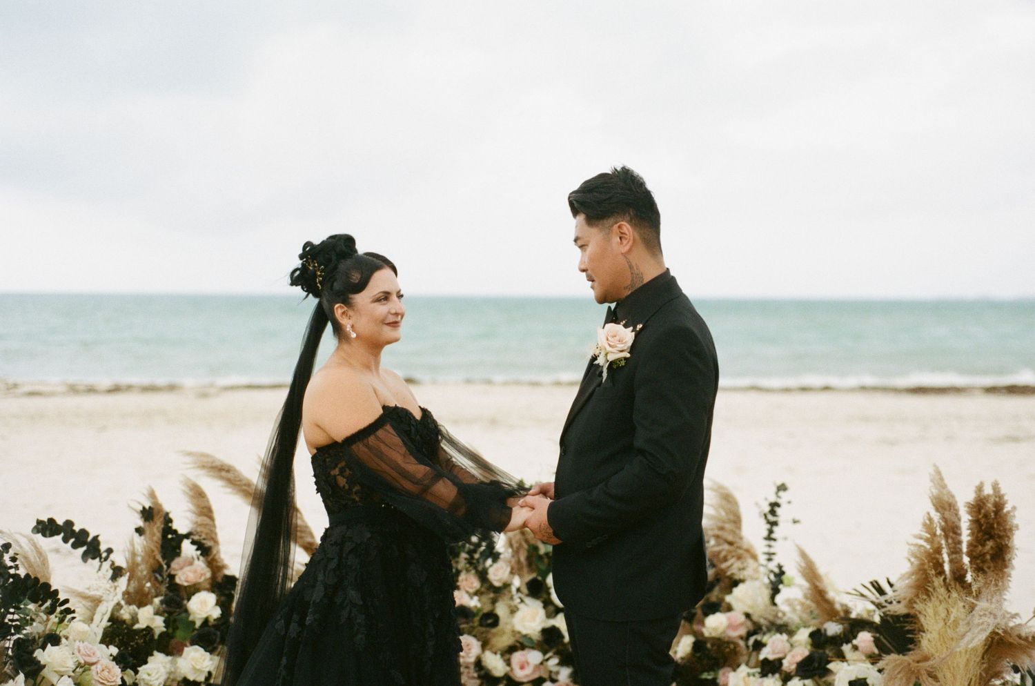 Bride in black lace gown with long veil exchanging vows with groom at beach ceremony with white florals.