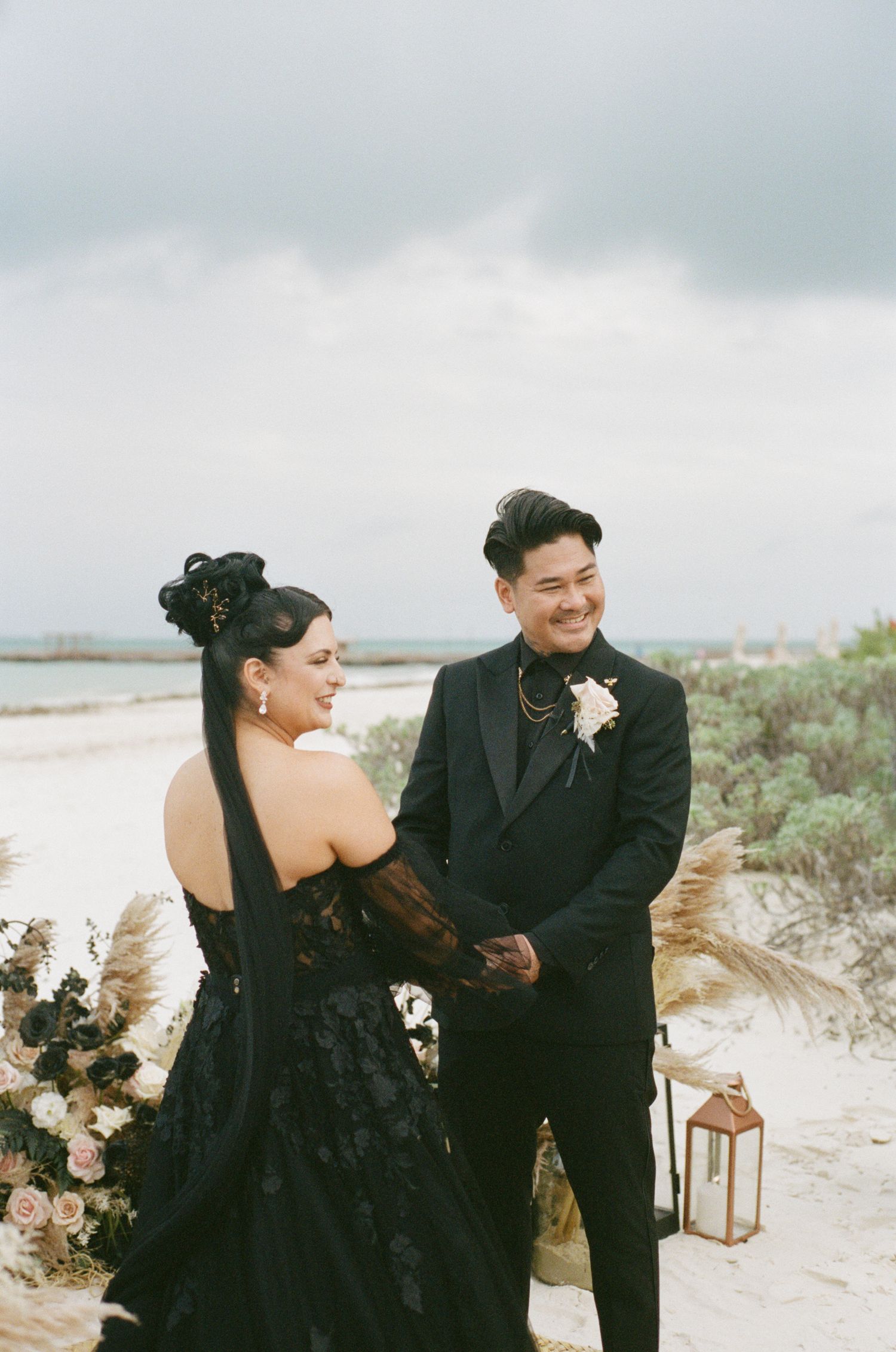Smiling groom in black tuxedo holds brides hands during beach wedding ceremony with pampas grass and rose decor.