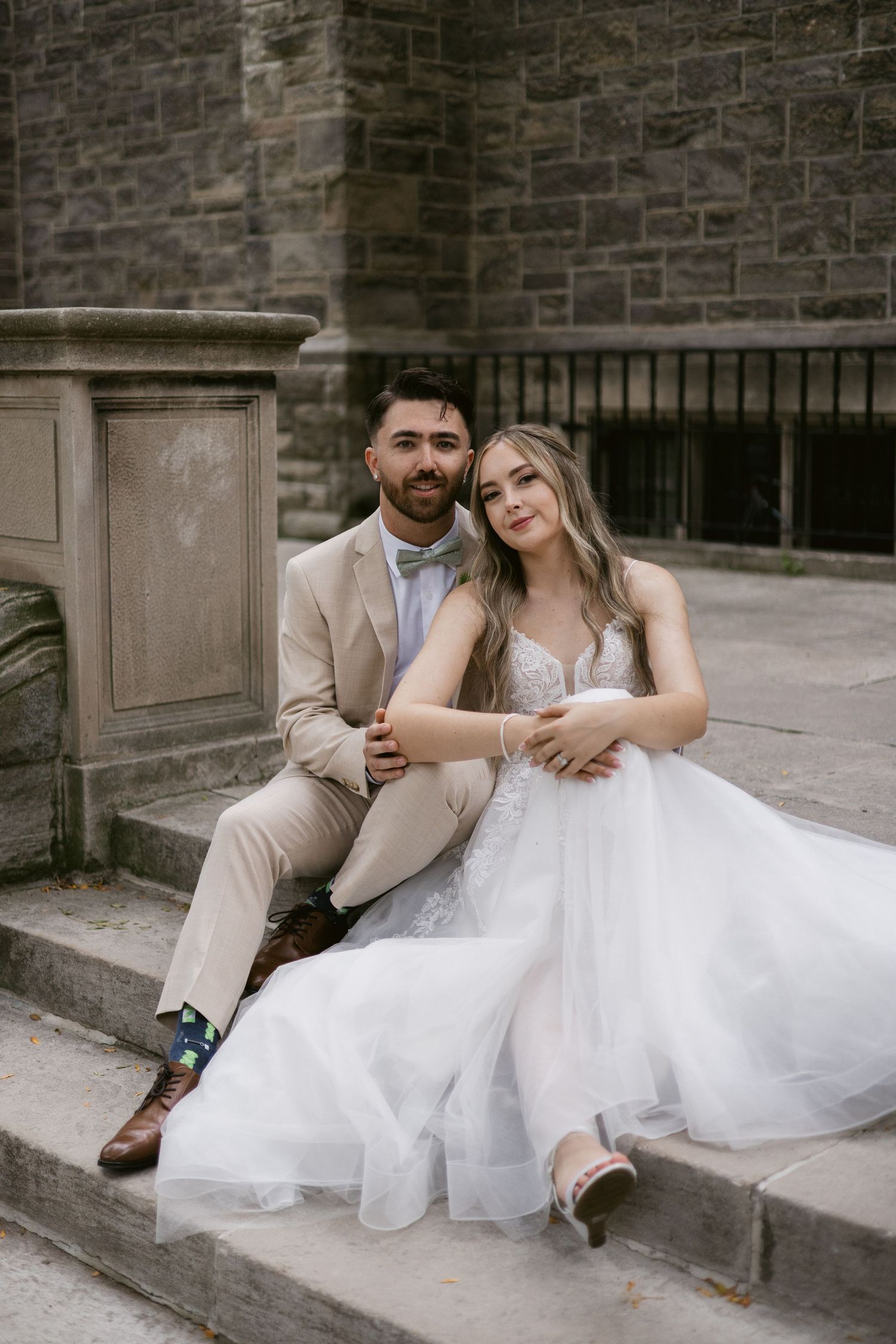 Bride in white tulle gown and groom in beige suit sit together on stone steps at a castle.