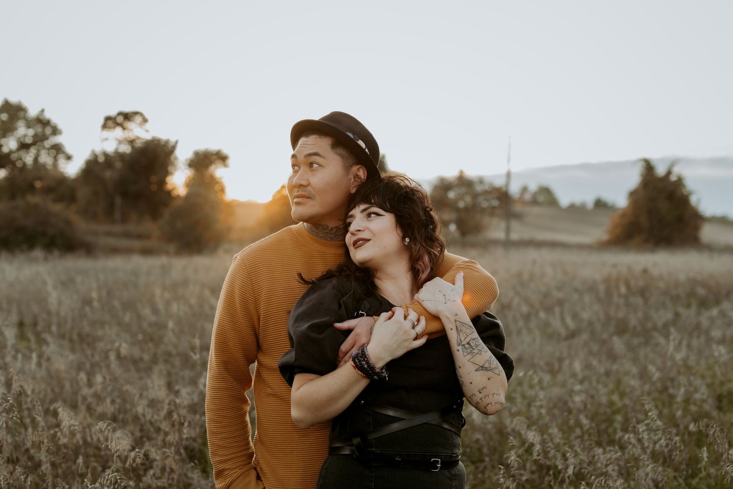 Couple embracing in golden sunset field, woman with tattoos leaning into man wearing mustard sweater and black hat.