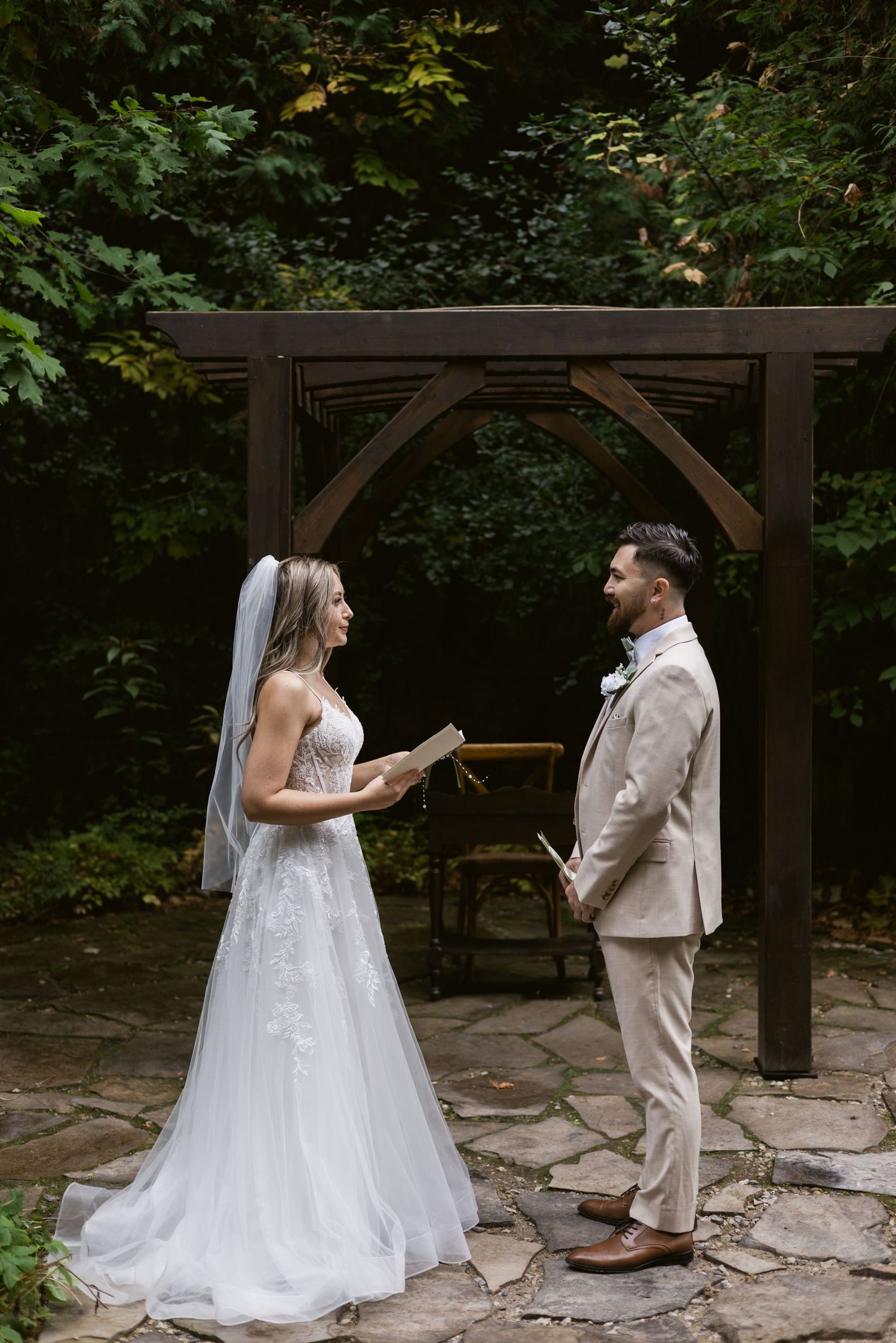 Bride and groom exchanging vows under wooden pergola arch surrounded by green trees outdoors.