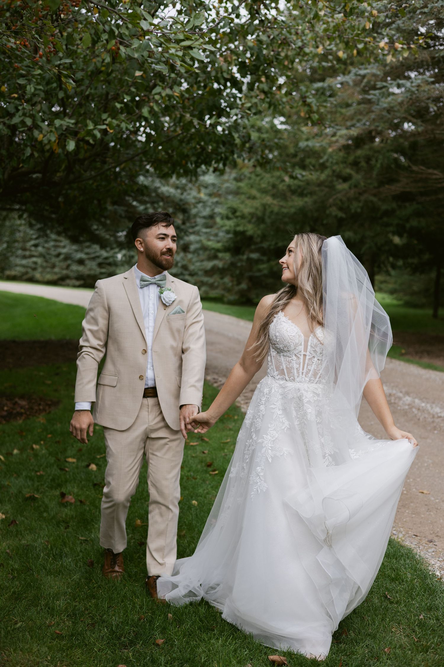 Bride and groom walk hand in hand through lush green garden, bride in lace gown with veil, groom in tan suit.