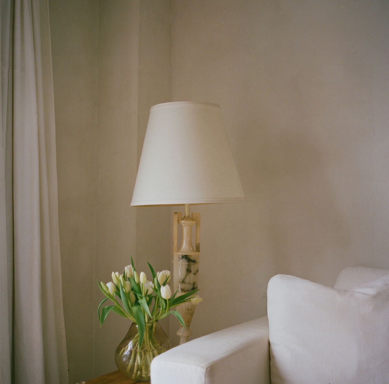 White tulips in glass vase beside a table lamp with cream shade in a soft neutral living room corner.