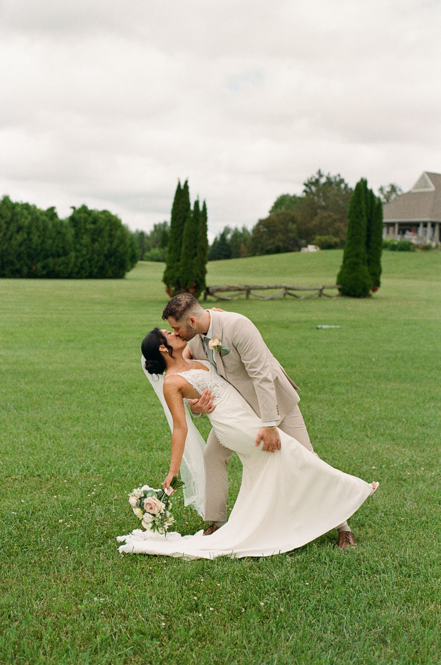Groom dips bride for a kiss on lush green lawn, bride holding floral bouquet in elegant white gown and veil.