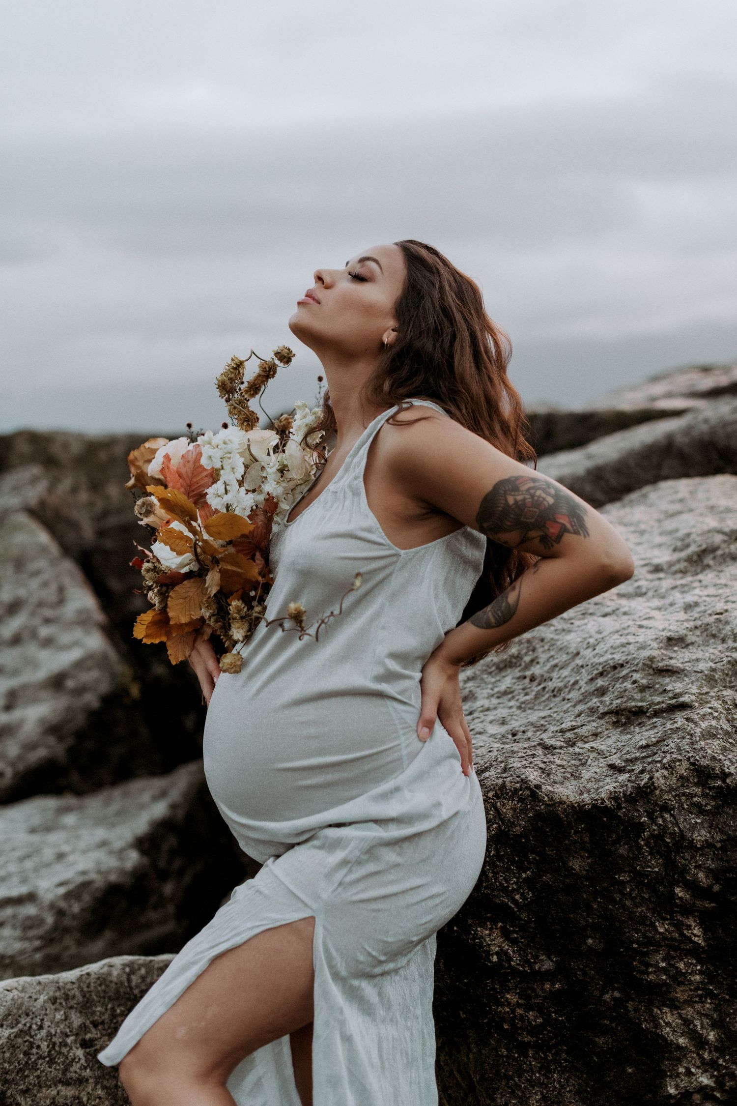 Pregnant woman in white dress holding autumn bouquet poses against rocks outdoors.
