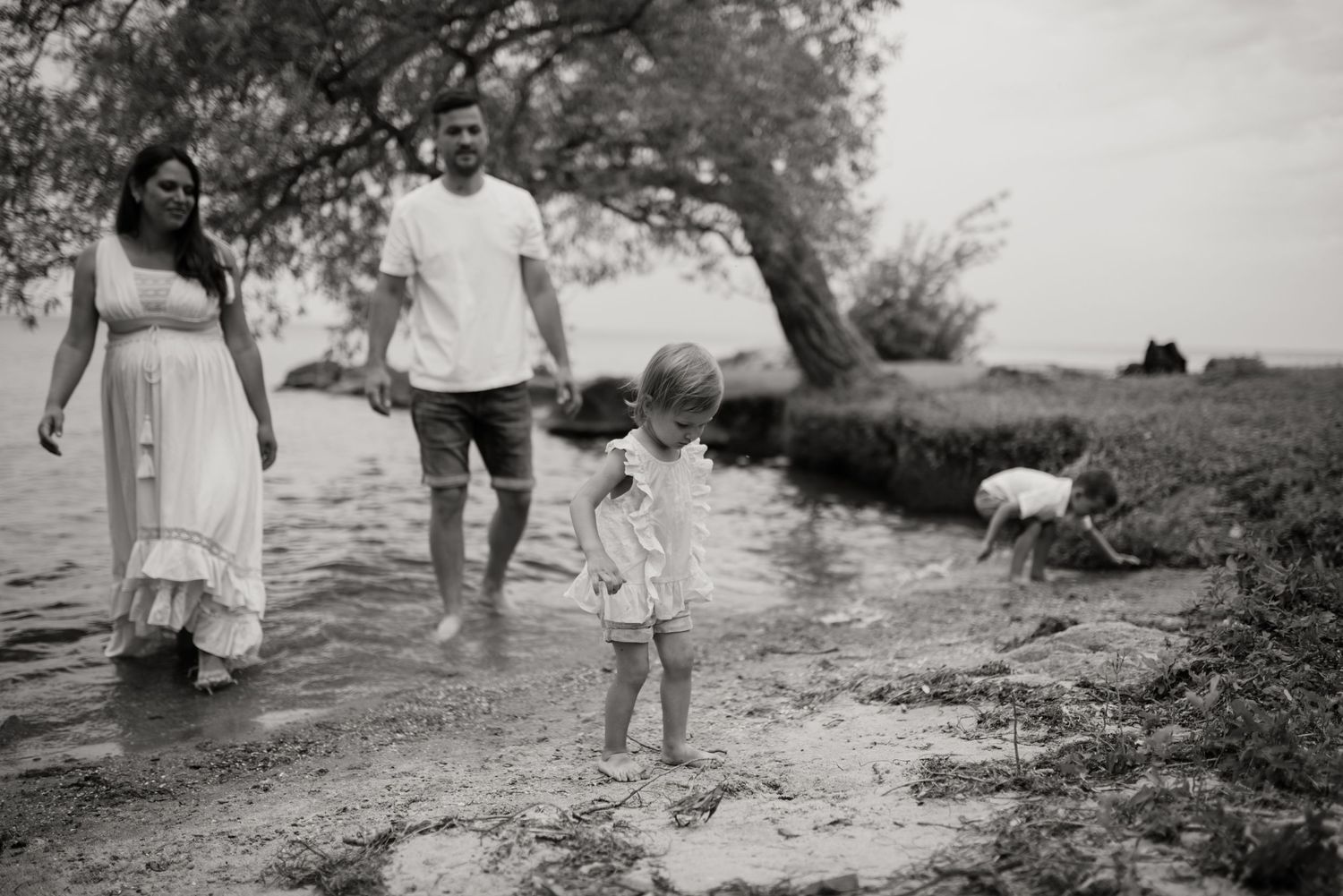 Black and white photo of expecting couple and toddler walking along a lakeside shore.