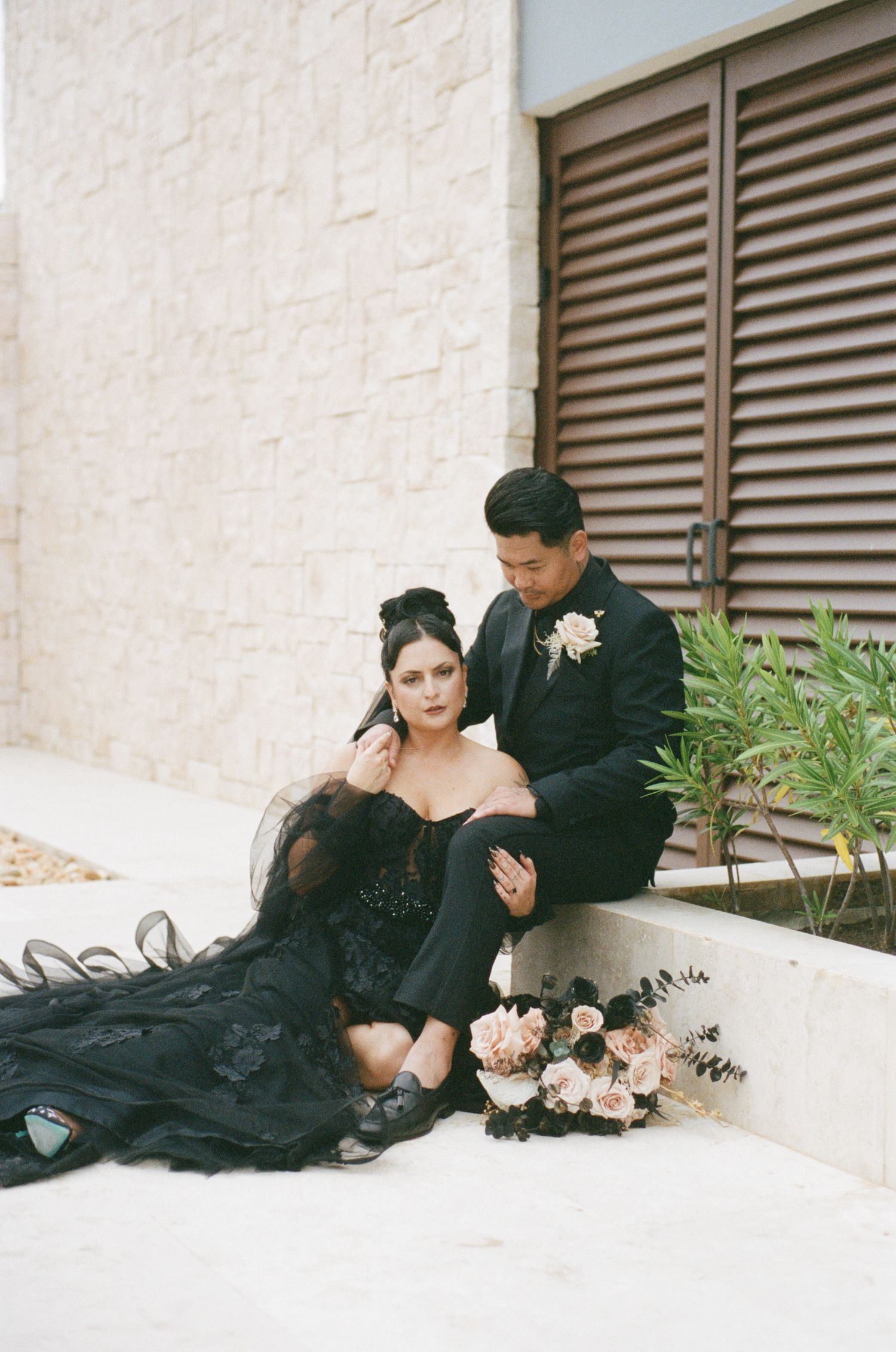 Bride in black gown and groom sit together against stone wall with bouquet.