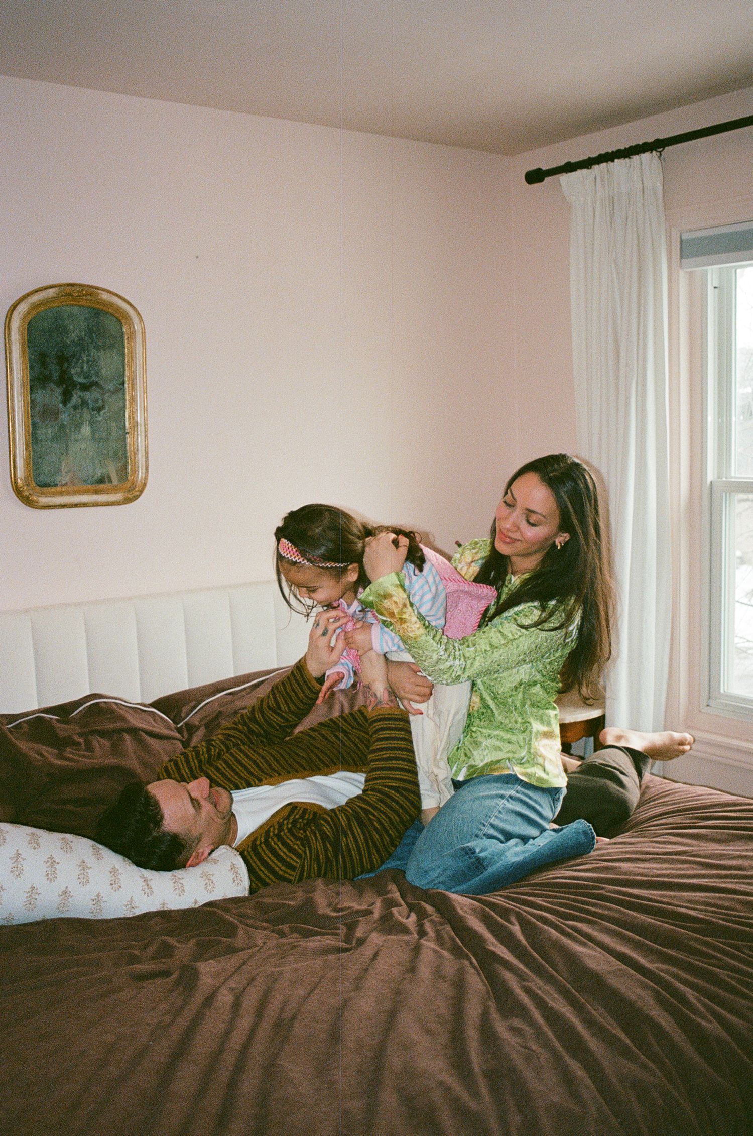 Woman in green top braids young girls hair on brown bed while man rests with pillow nearby.