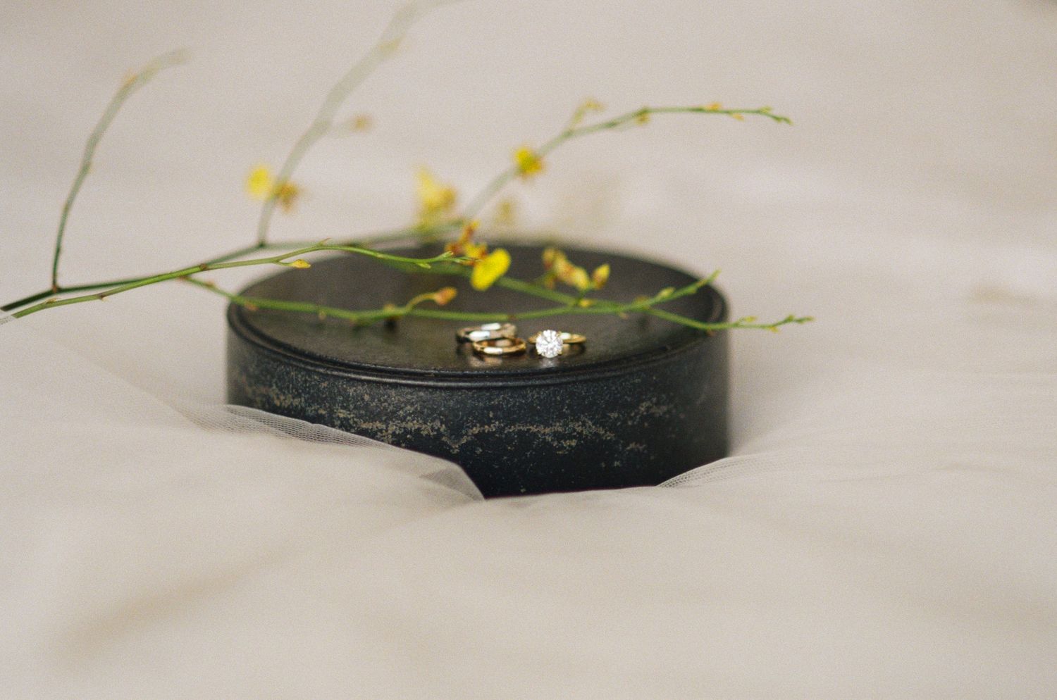 Gold wedding rings rest on a dark decorative dish with delicate yellow flowers on white fabric.