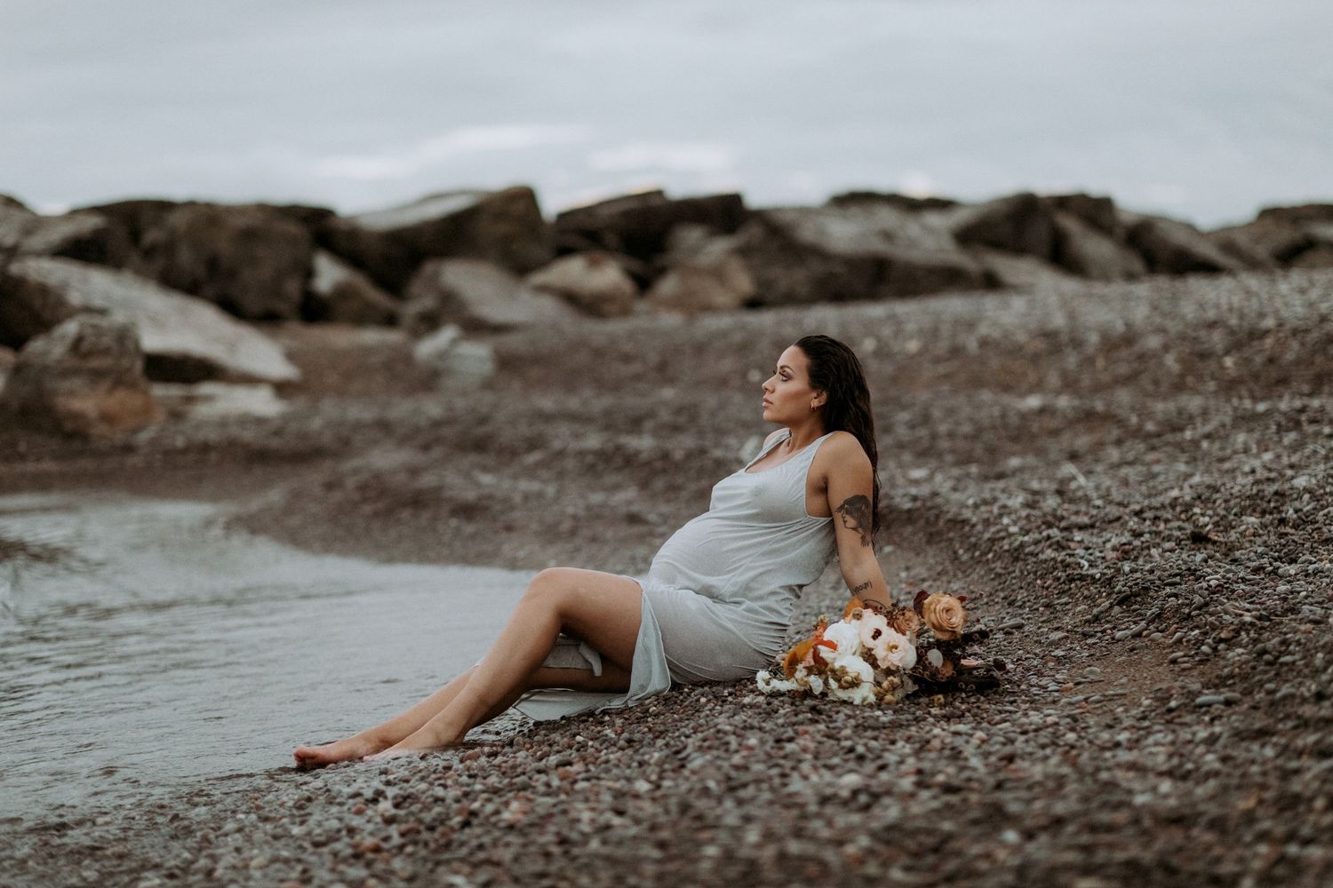 Pregnant woman in grey dress sits on rocky shoreline, gazing away with bouquet of flowers beside her.