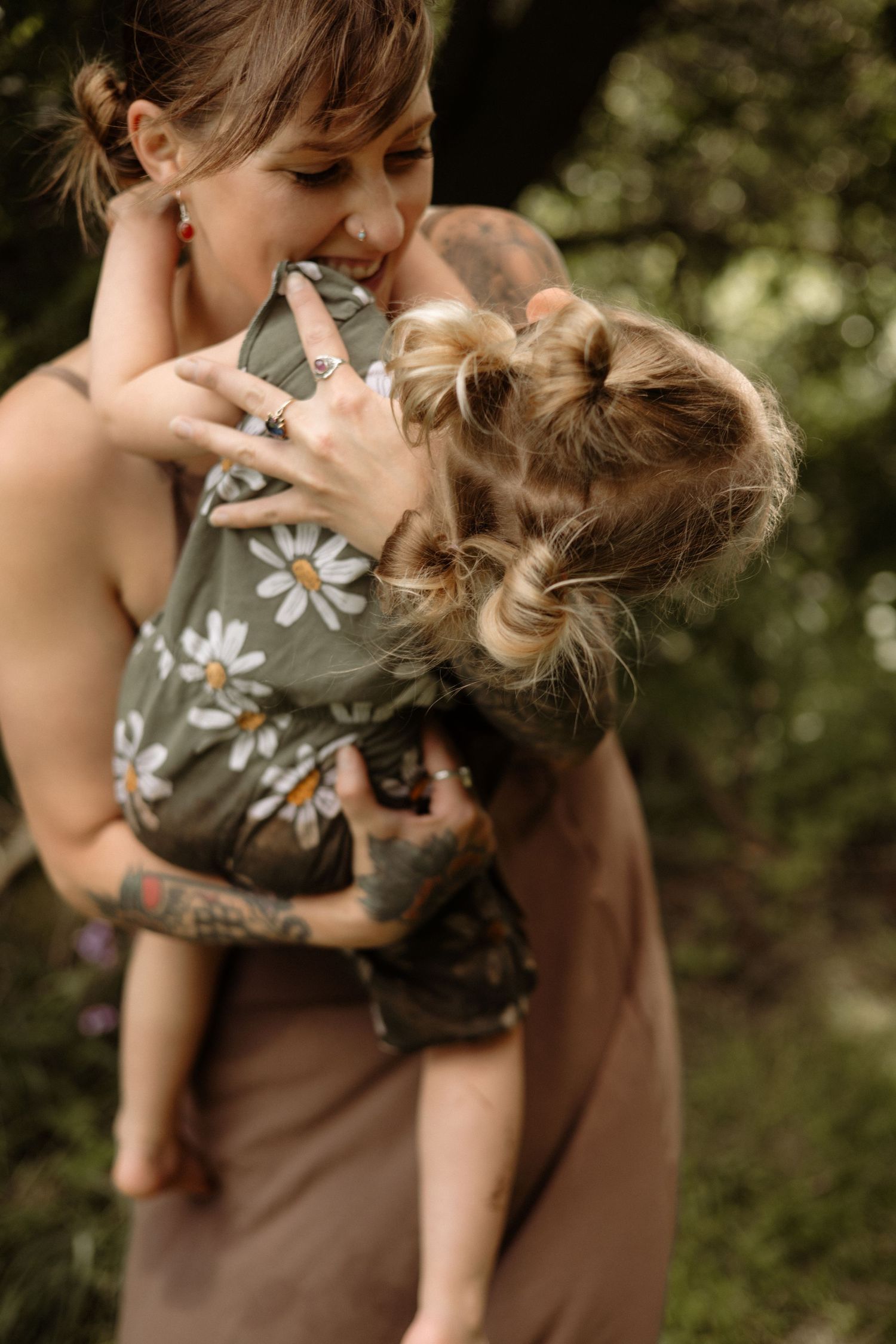 A smiling tattooed mother holds her laughing young child in a garden setting.