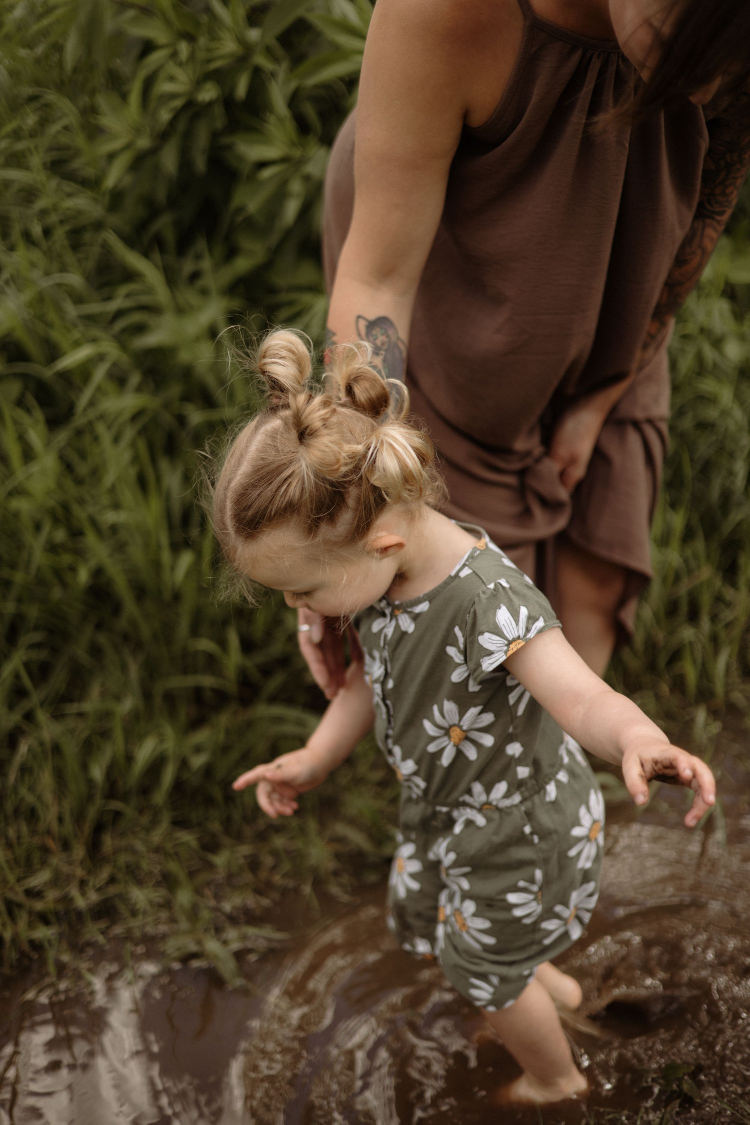 A tattooed mother guides her toddler girl in a daisy romper through a muddy puddle outdoors.