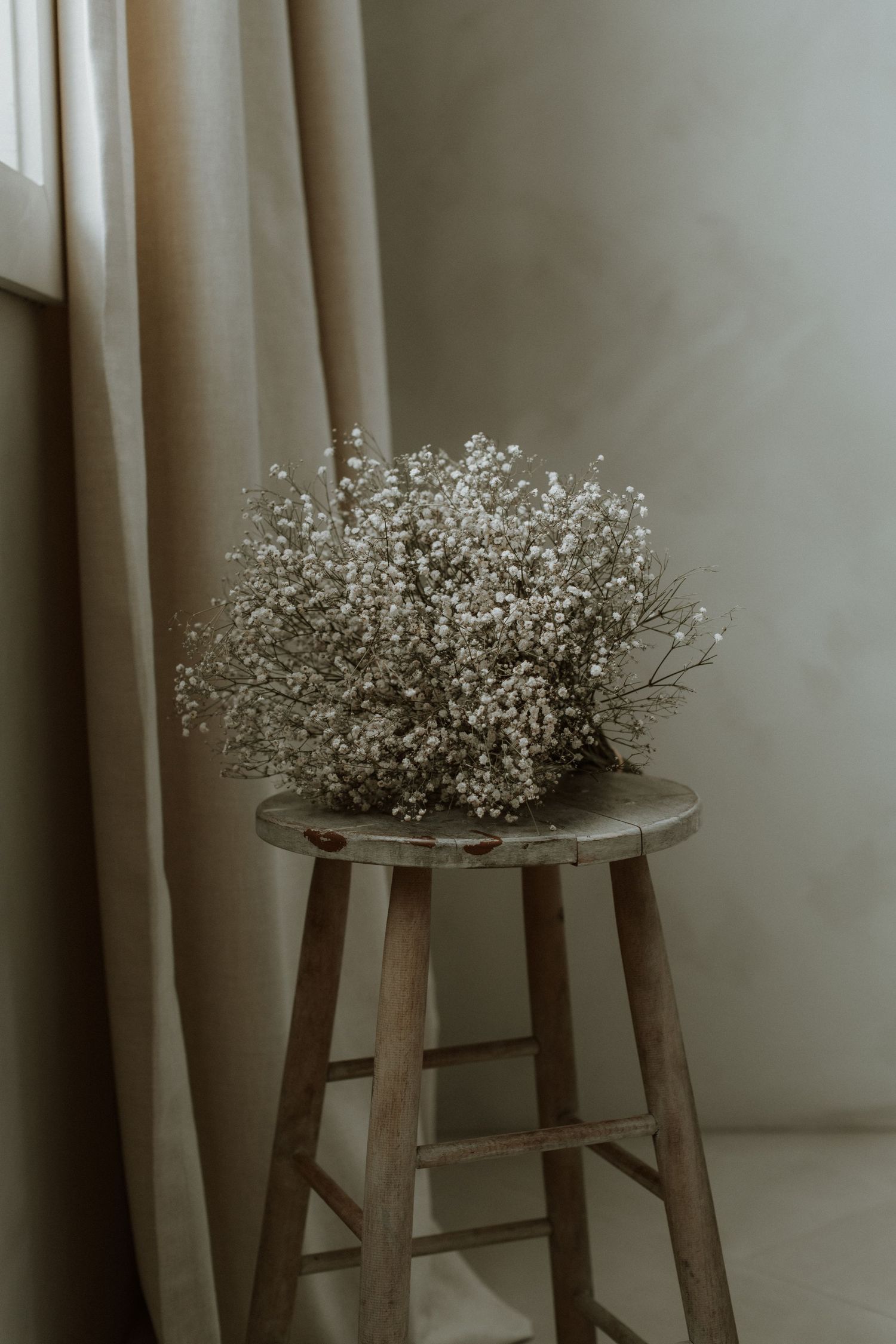 Bouquet of white baby's breath flowers resting on a rustic wooden stool near a linen curtain.