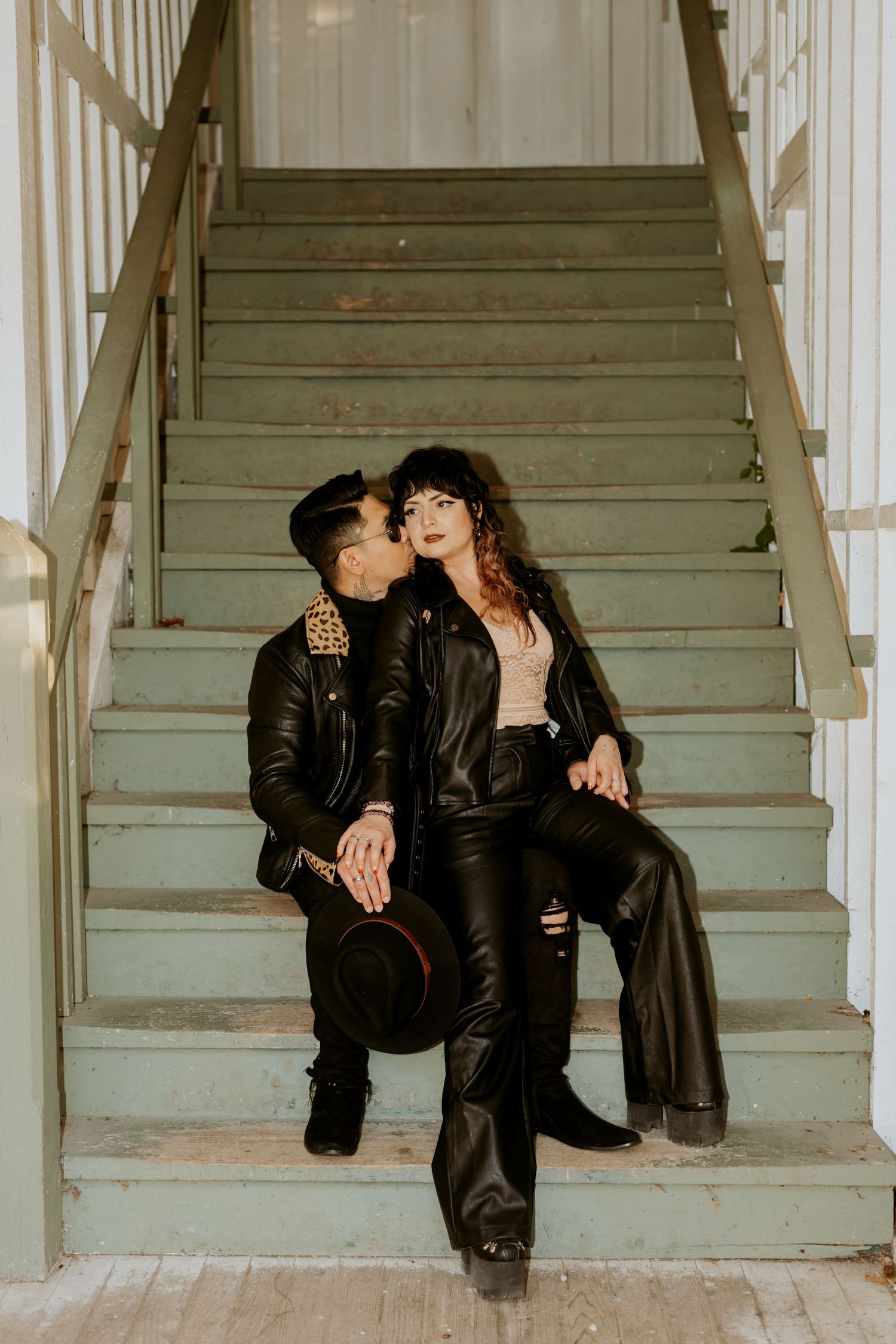 Man kissing woman on cheek while sitting on vintage green stairs in leather jackets.