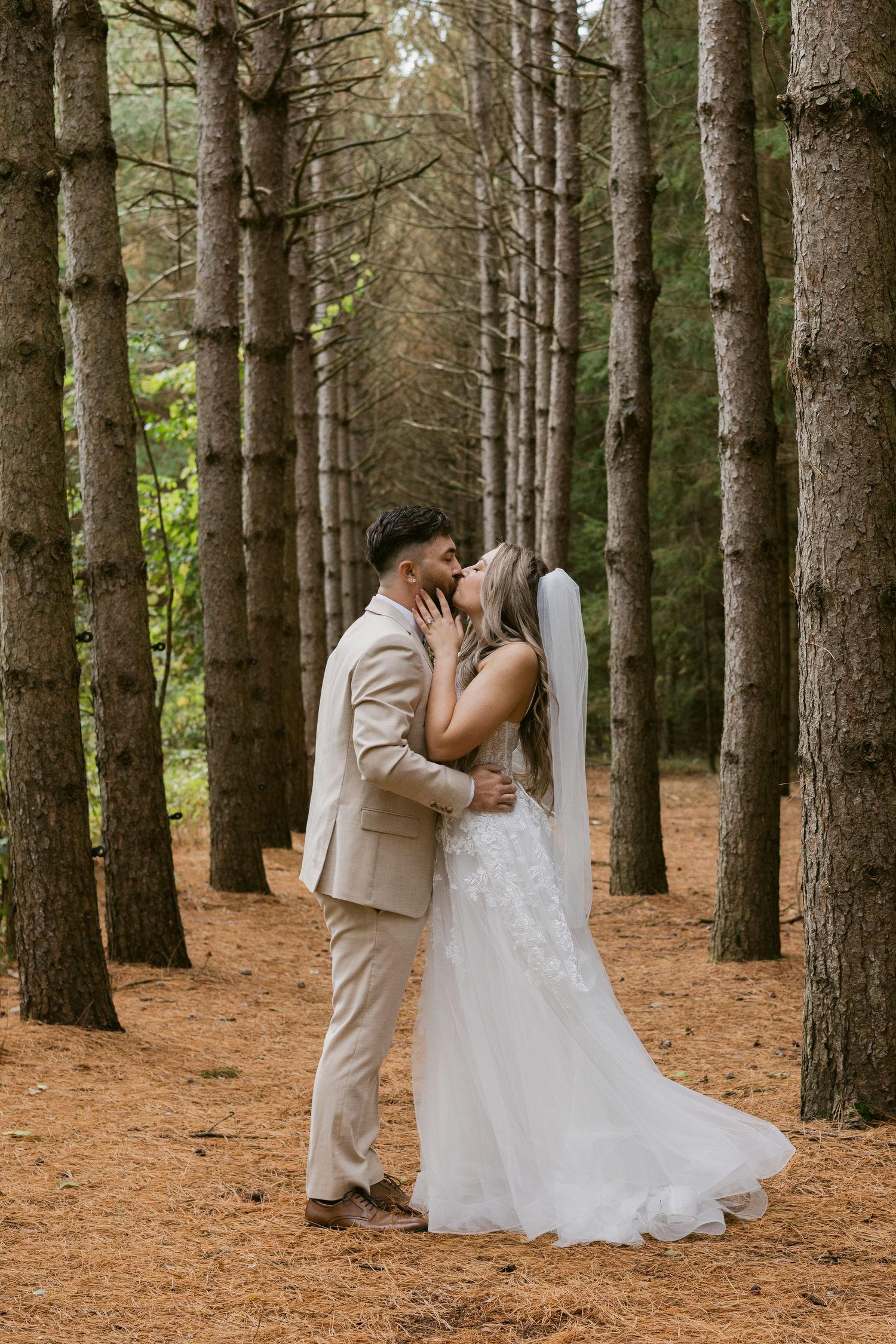 Bride and groom share a romantic kiss surrounded by tall pine trees on their wedding day.