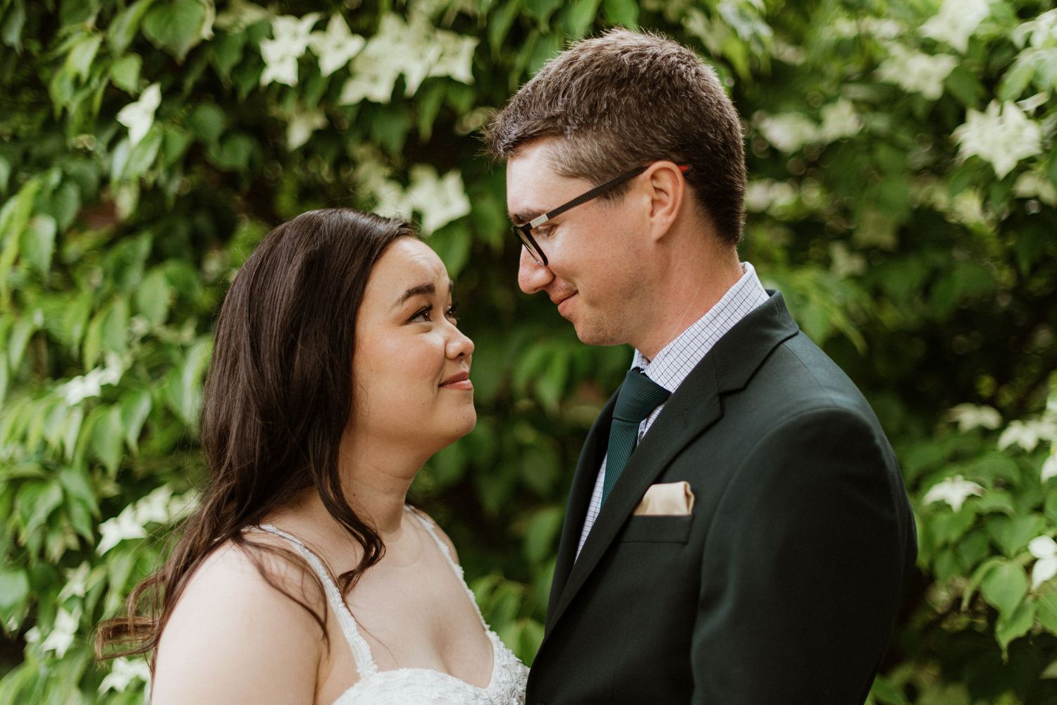 Bride and groom share an intimate glance surrounded by lush green foliage on wedding day.