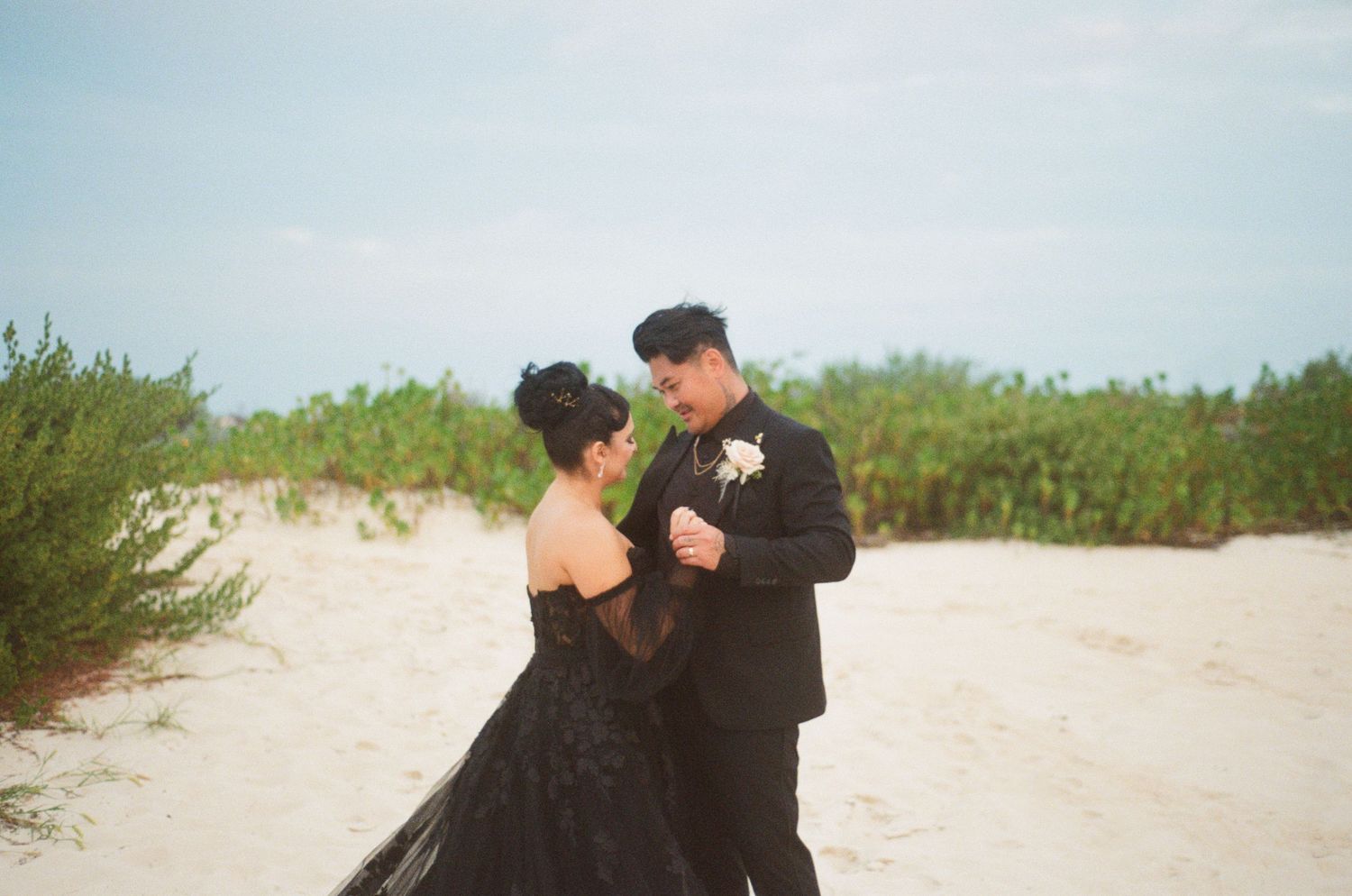 Bride and groom in black wedding attire share romantic dance on sandy beach.