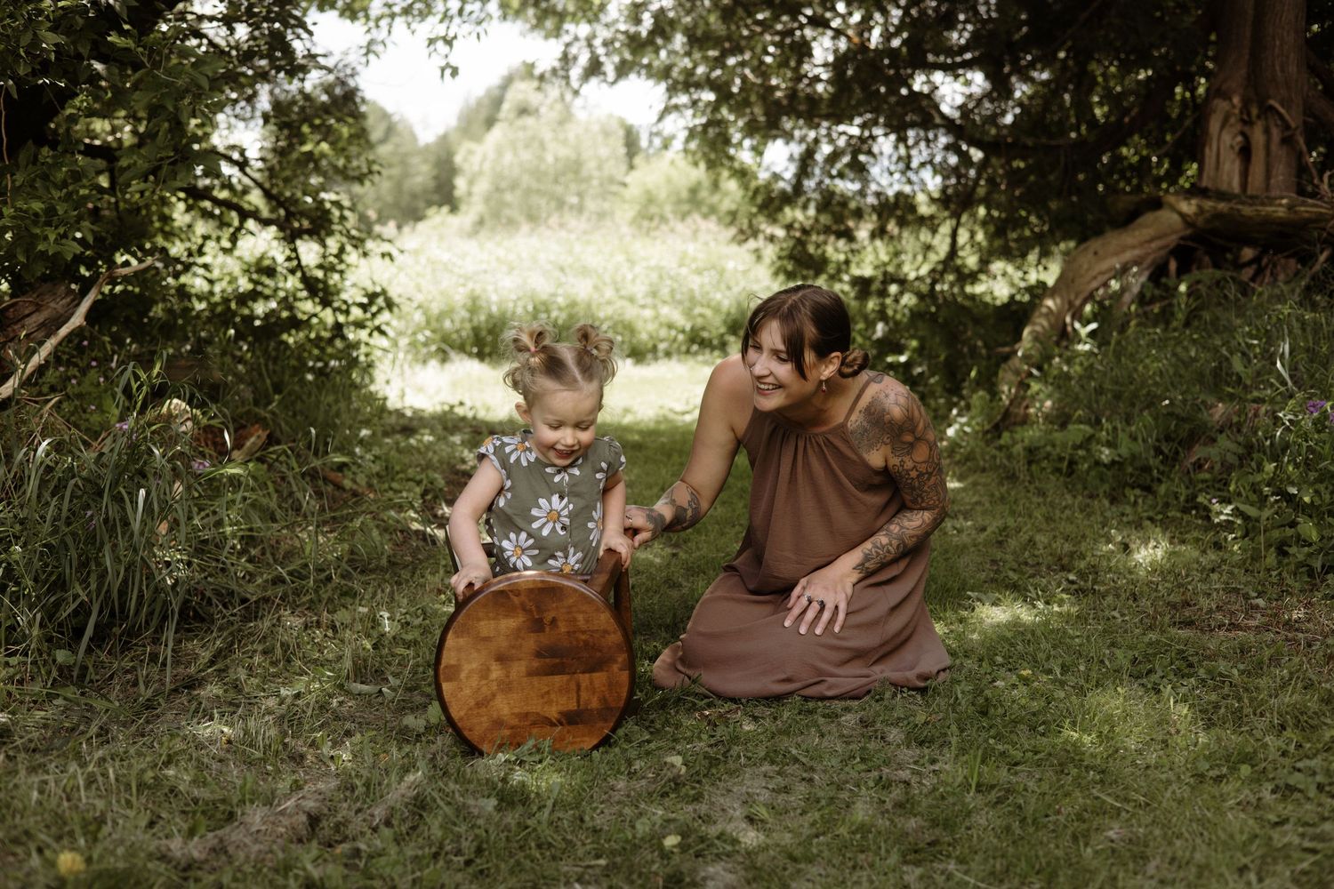 Tattooed mother and toddler daughter sit together on grass exploring a round wooden object under leafy trees.