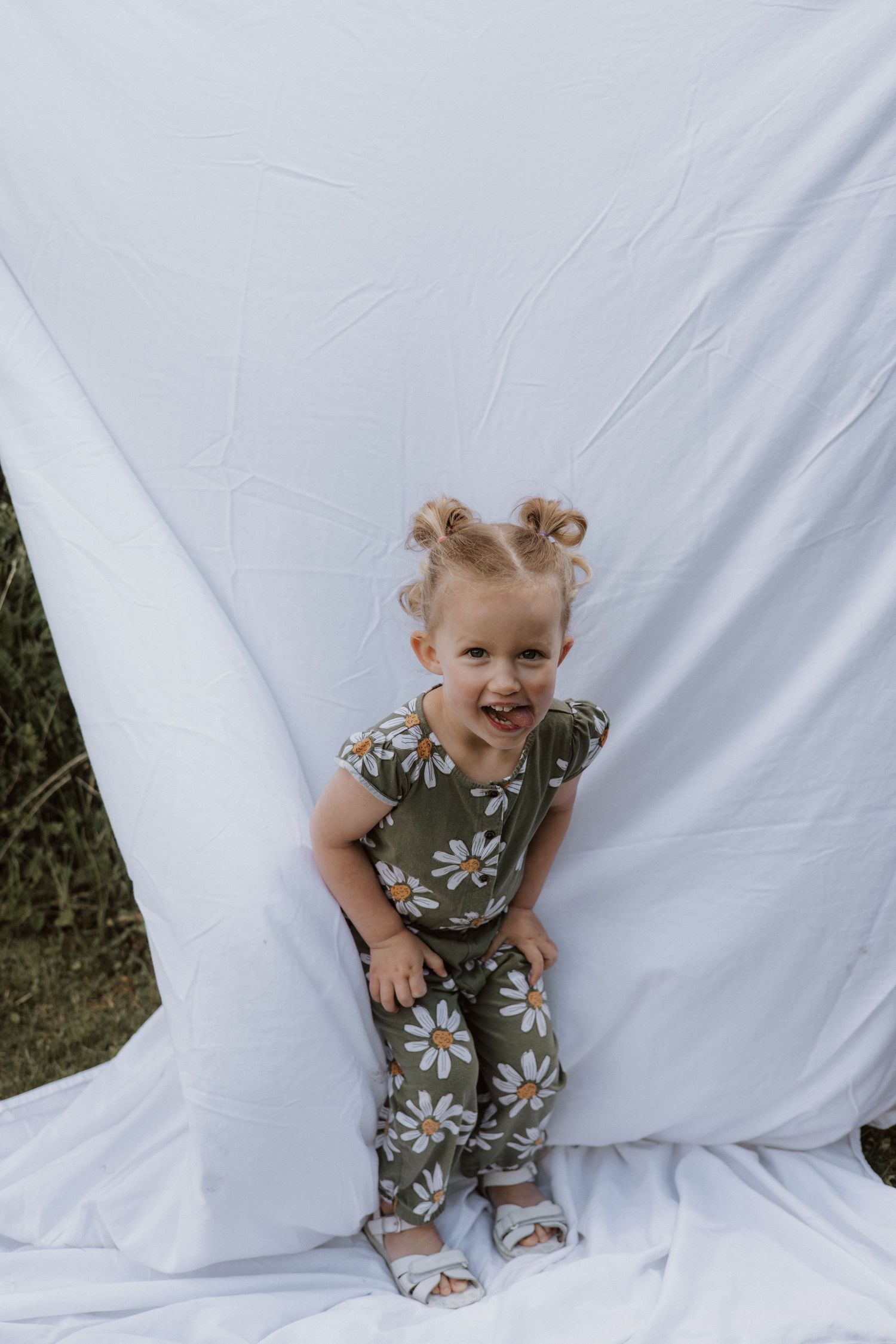 Cheerful toddler girl in daisy floral outfit with pigtail buns posing playfully against a white fabric backdrop.