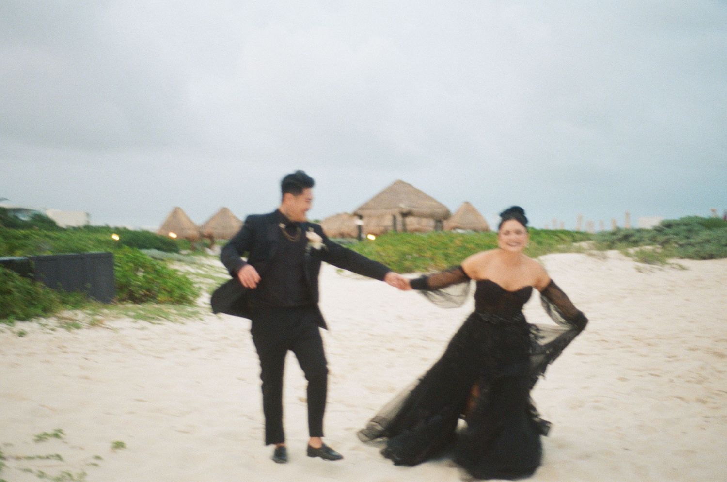 Bride in black lace gown and groom in black suit holding hands on a white sand beach.