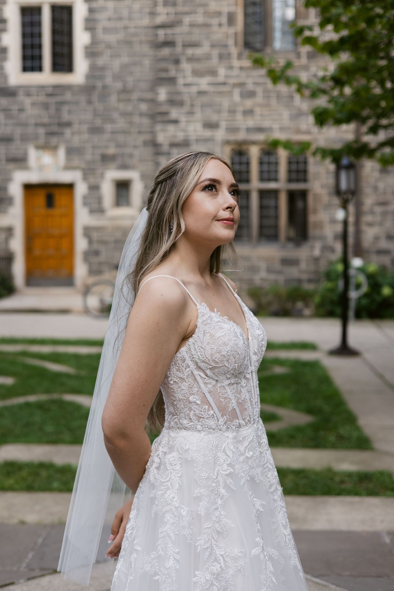 Bride in a lace A-line wedding gown with veil stands before a stone building, looking elegantly to the side.