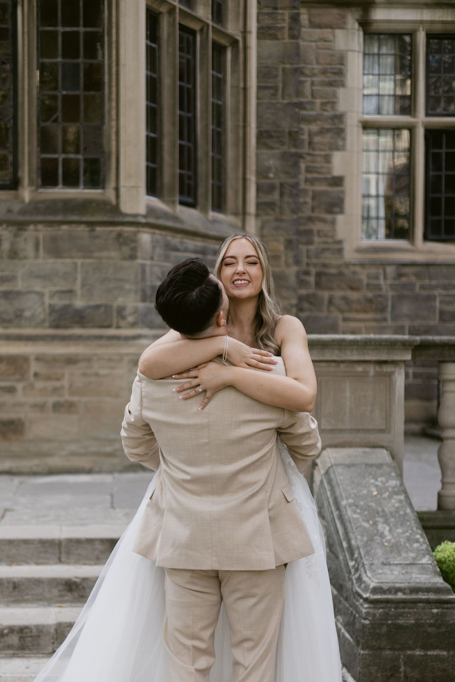 Bride laughs joyfully while embracing groom outside a historic stone building on their wedding day.