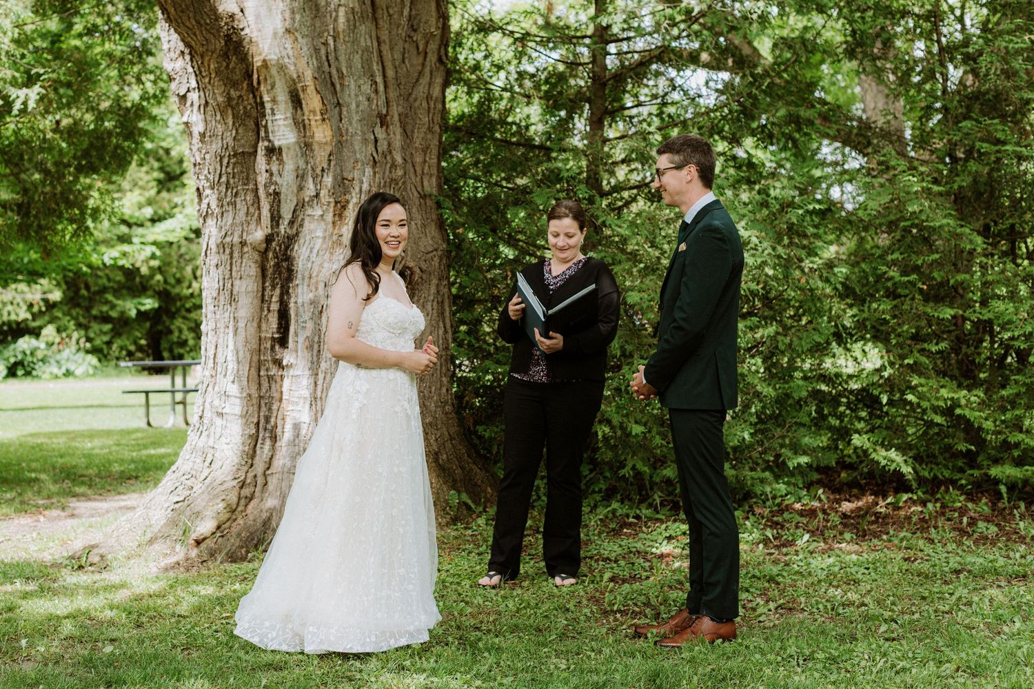 Bride and groom exchange vows with officiant beside a large tree in a lush outdoor ceremony.