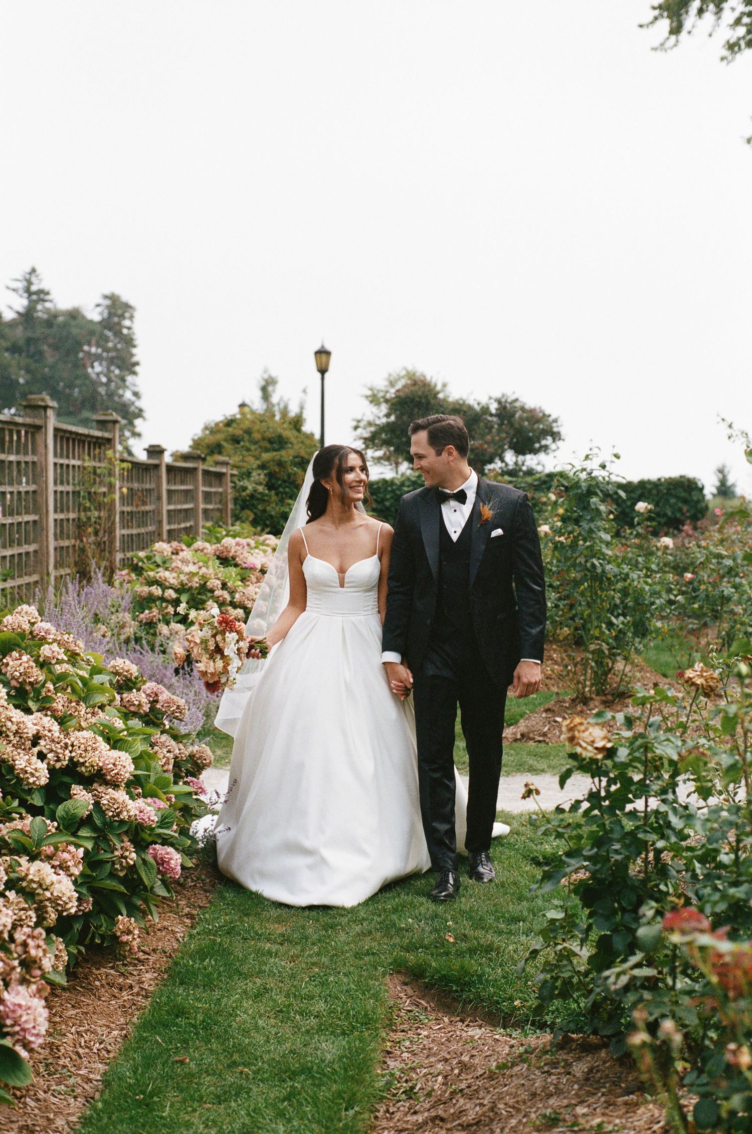 Bride in white ballgown and groom in black tuxedo walk hand in hand through a garden.