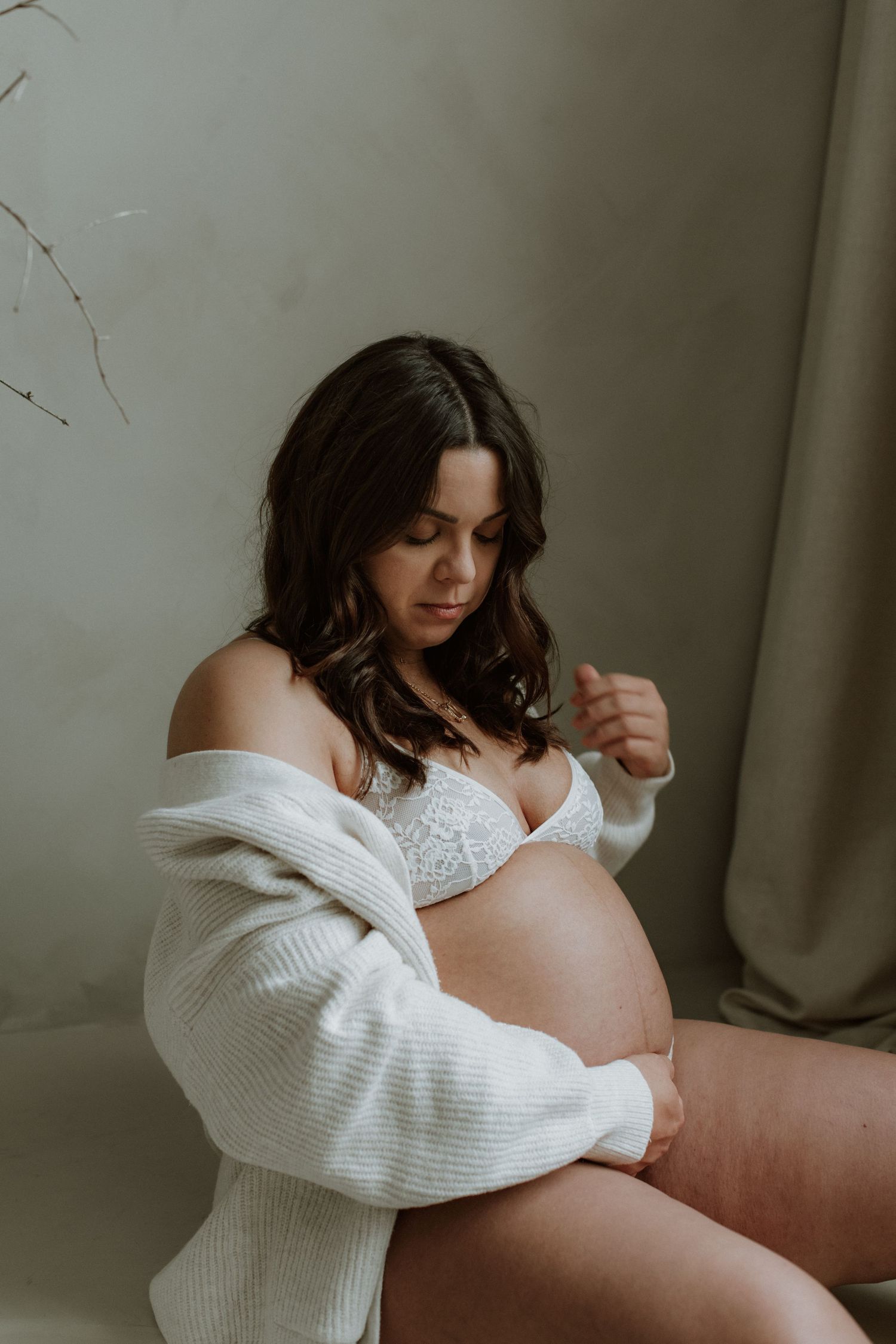 Pregnant woman in white lace lingerie and cardigan looks down at her baby bump in a studio.