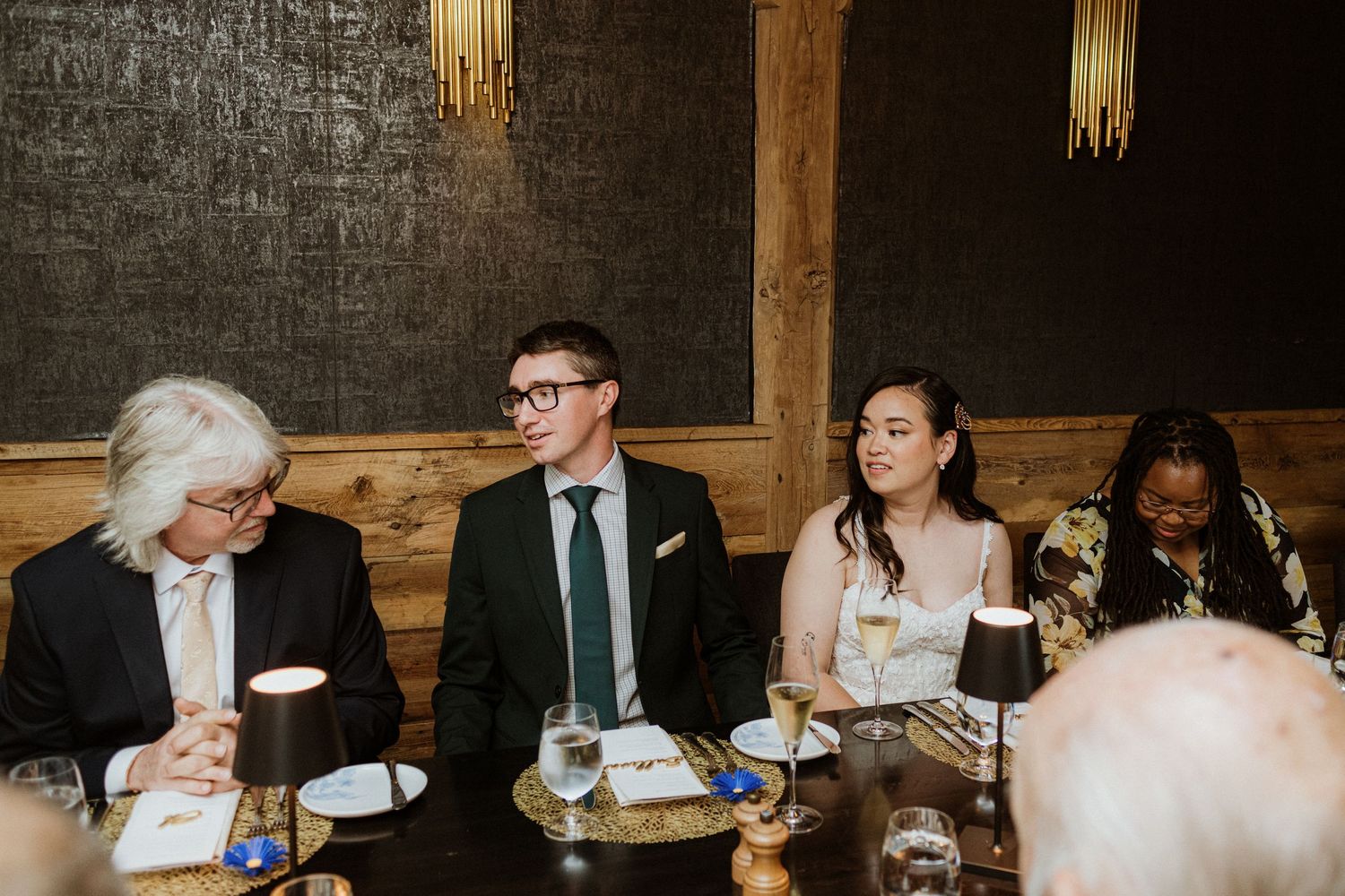Bride and groom seated with guests at wedding reception dinner table in a dark wood-paneled room.